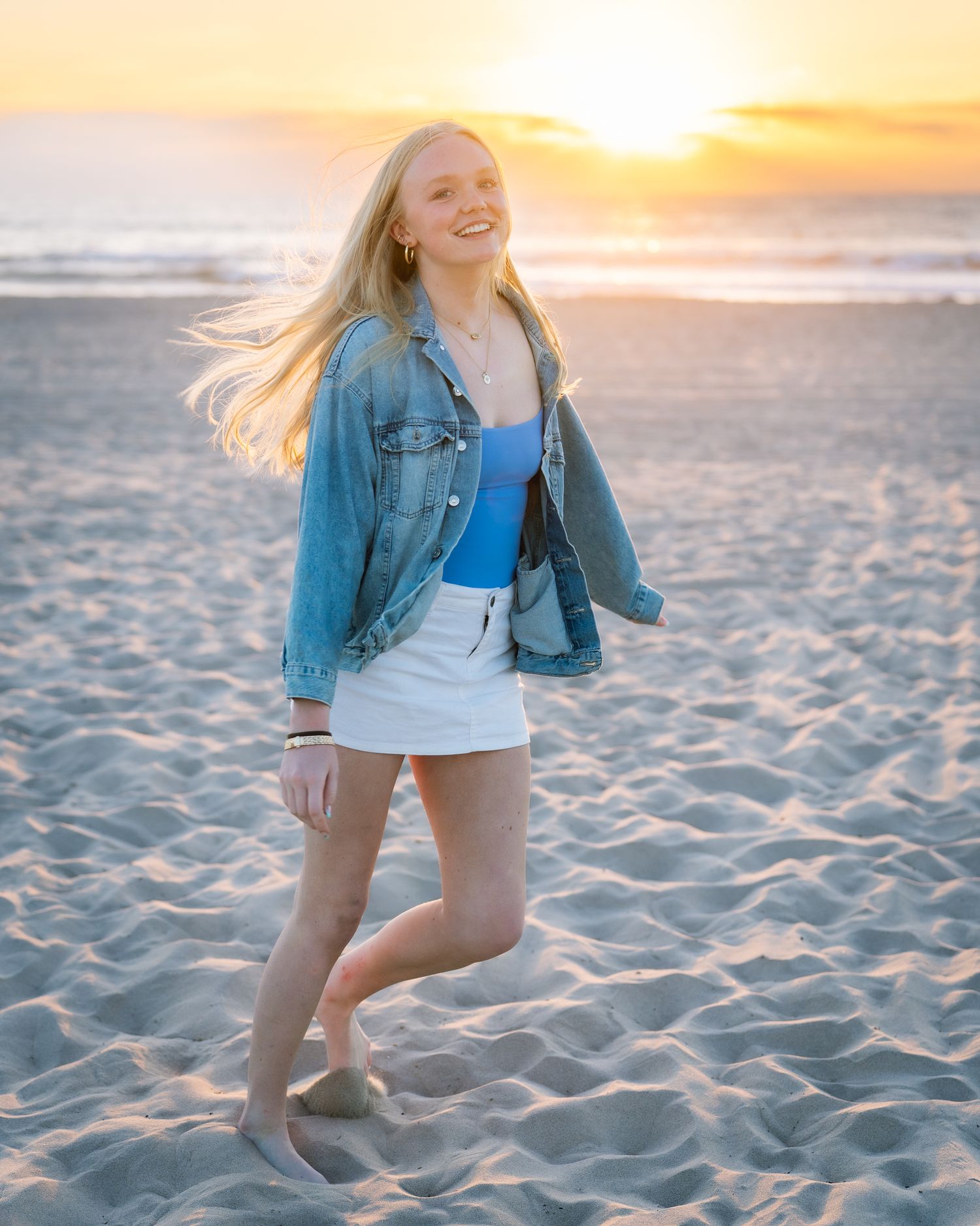 Smiling blonde woman in denim jacket and white skirt posing on sandy beach at golden sunset.