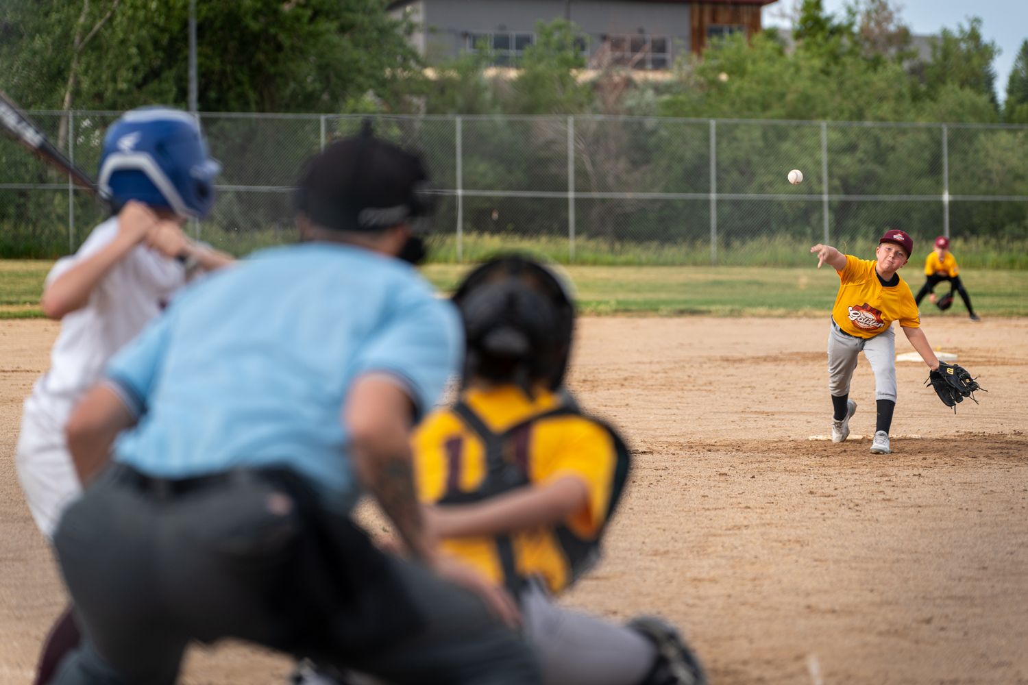 Baseball players compete in a recreational softball game on a dusty diamond field surrounded by chain link fences.
