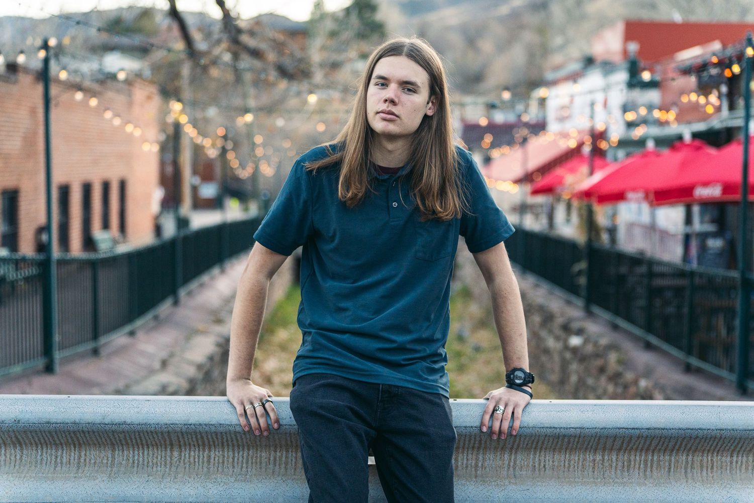 Young man with long hair in blue polo shirt leaning on railing in urban setting with string lights.