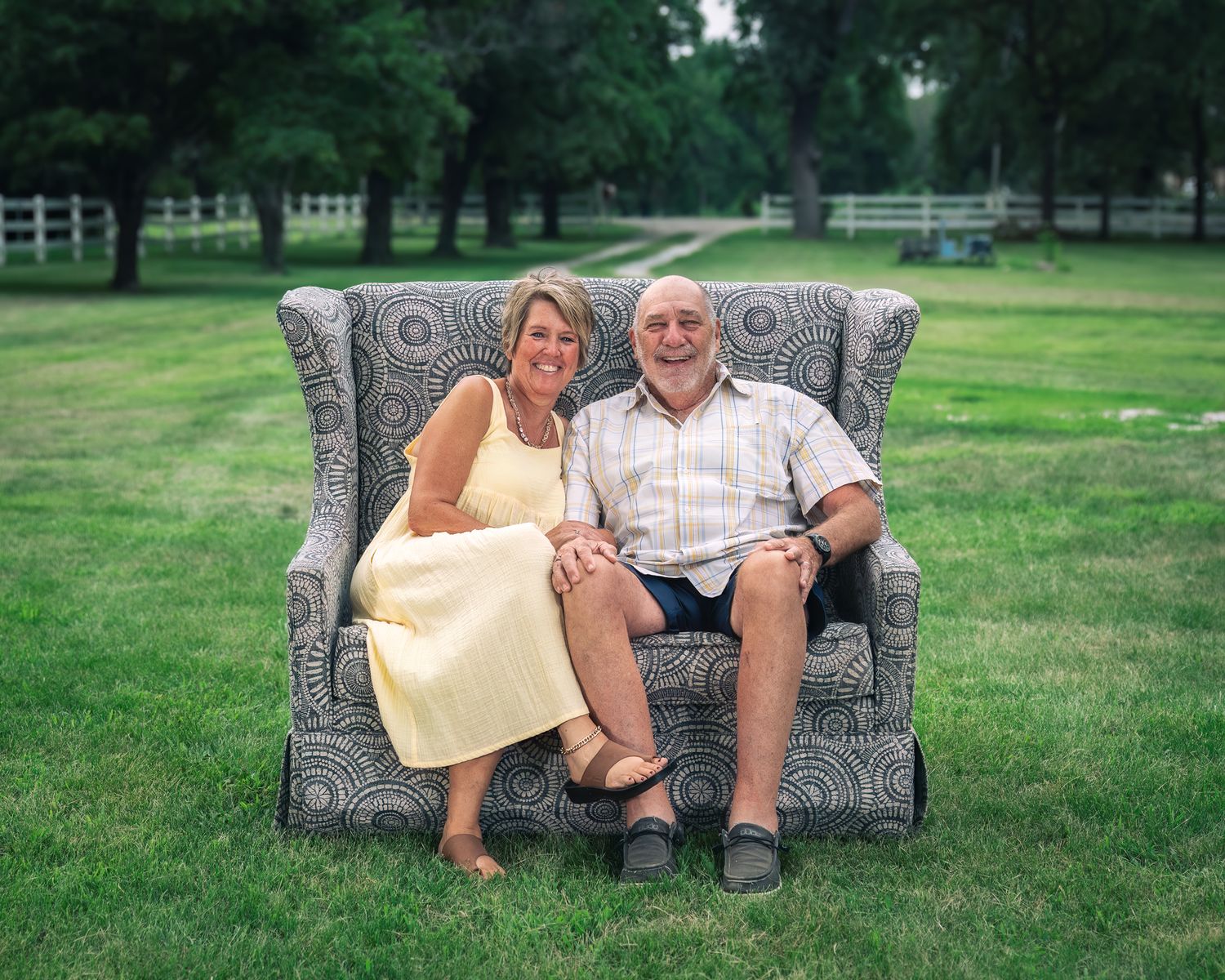 A couple sits together on a gray upholstered loveseat placed on a green lawn with trees in the background.