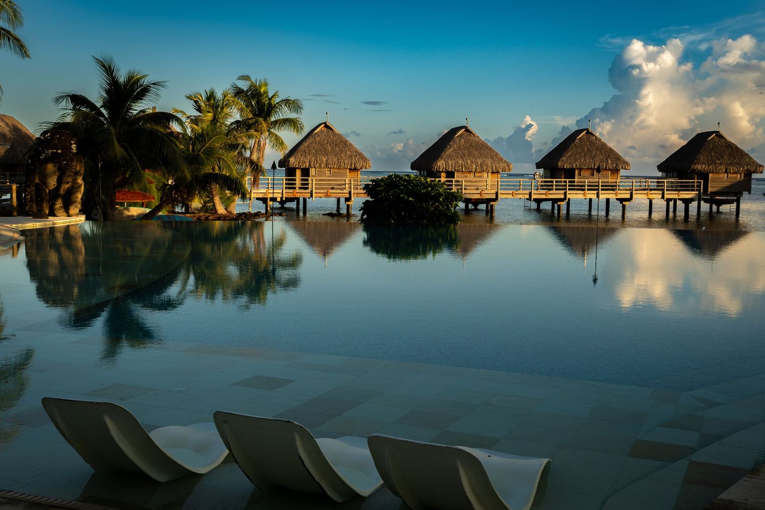 Tropical overwater bungalows reflect in a serene infinity pool at dusk, with palm trees and lounge chairs at a luxury resort.