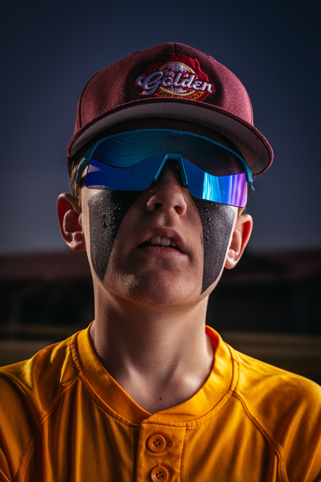Close-up portrait of a baseball player wearing a maroon cap with iridescent sunglasses at dusk, dressed in a yellow jersey.