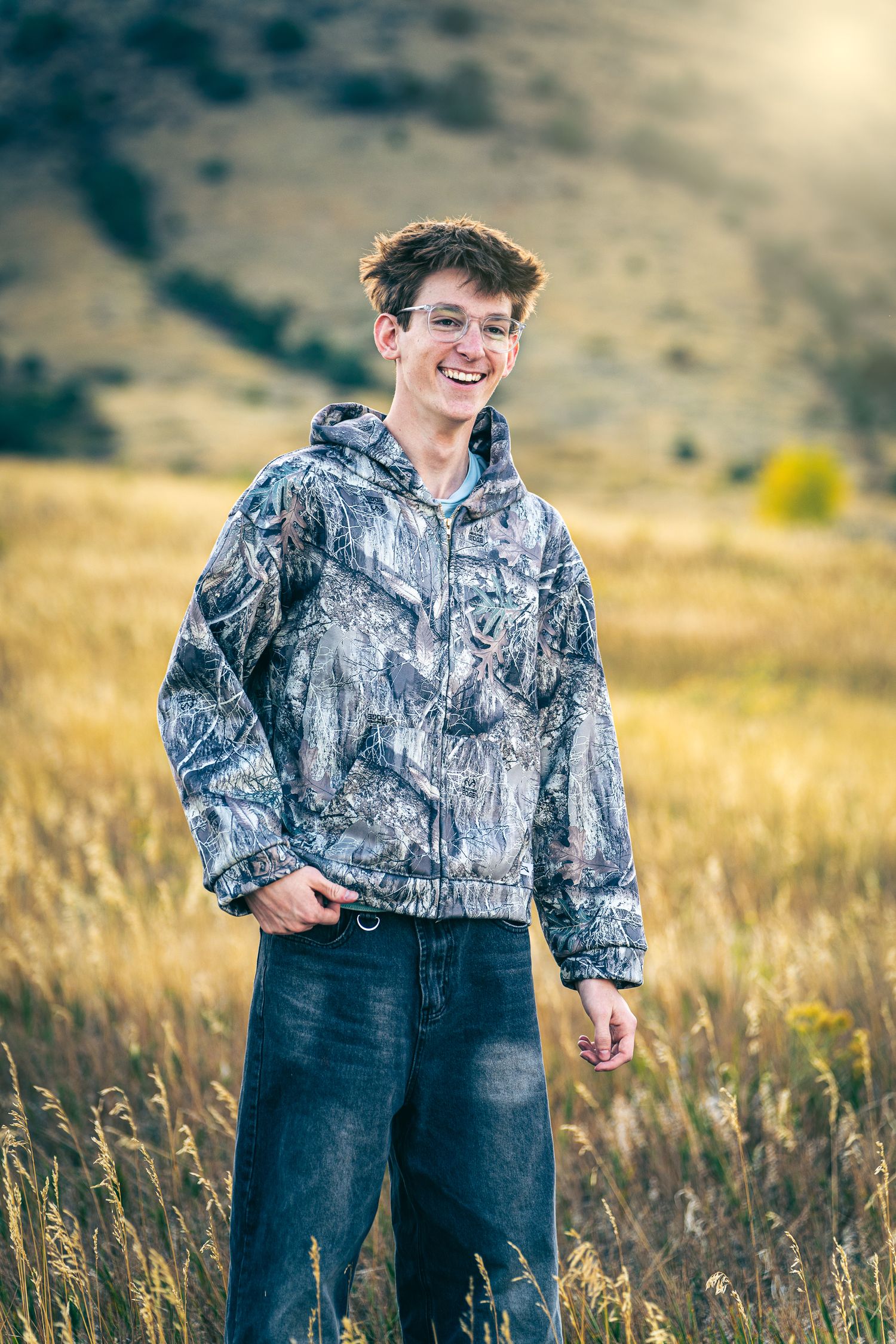 Smiling teenage boy in camo jacket and jeans standing in golden grass field with sunlit hills in background.