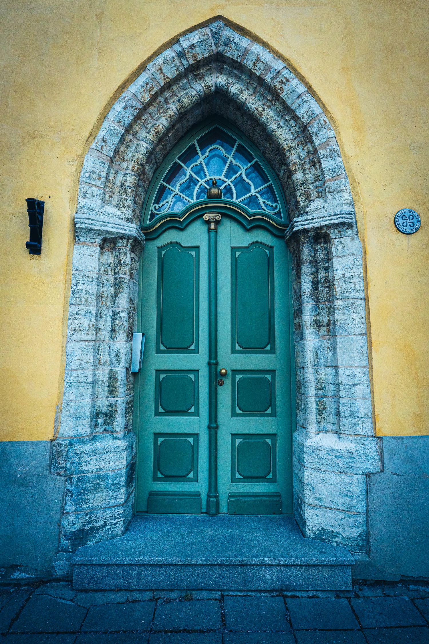 Ornate teal Gothic arched door with stained glass transom set in stone surround against yellow wall.