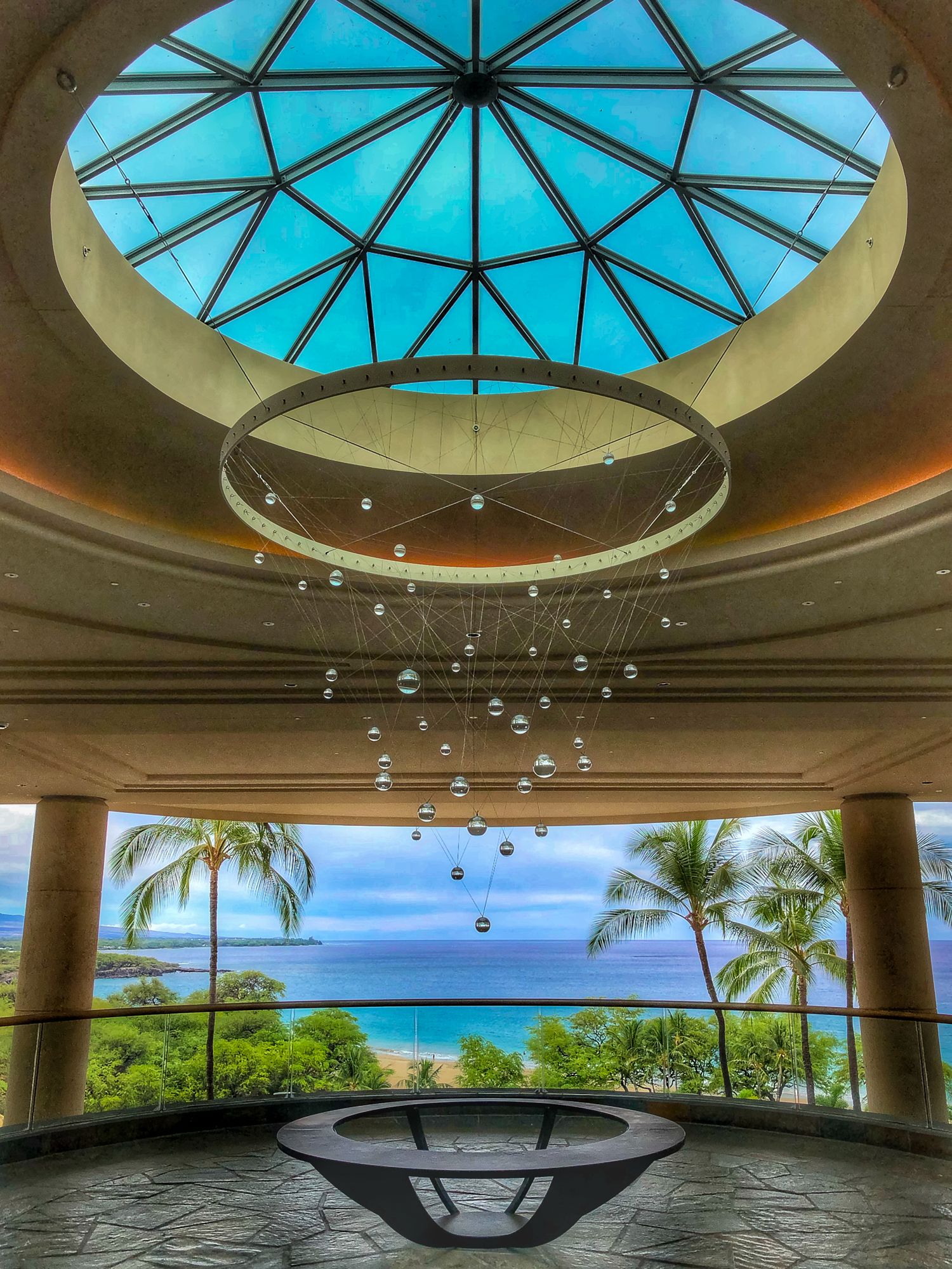 Luxury hotel lobby with geometric skylight, bubble chandelier, and stunning ocean view framed by tropical palm trees.