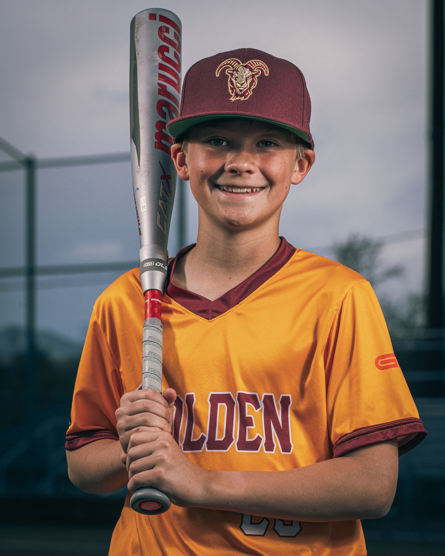 Baseball player in golden yellow uniform and maroon cap holds bat against blurred outdoor background.
