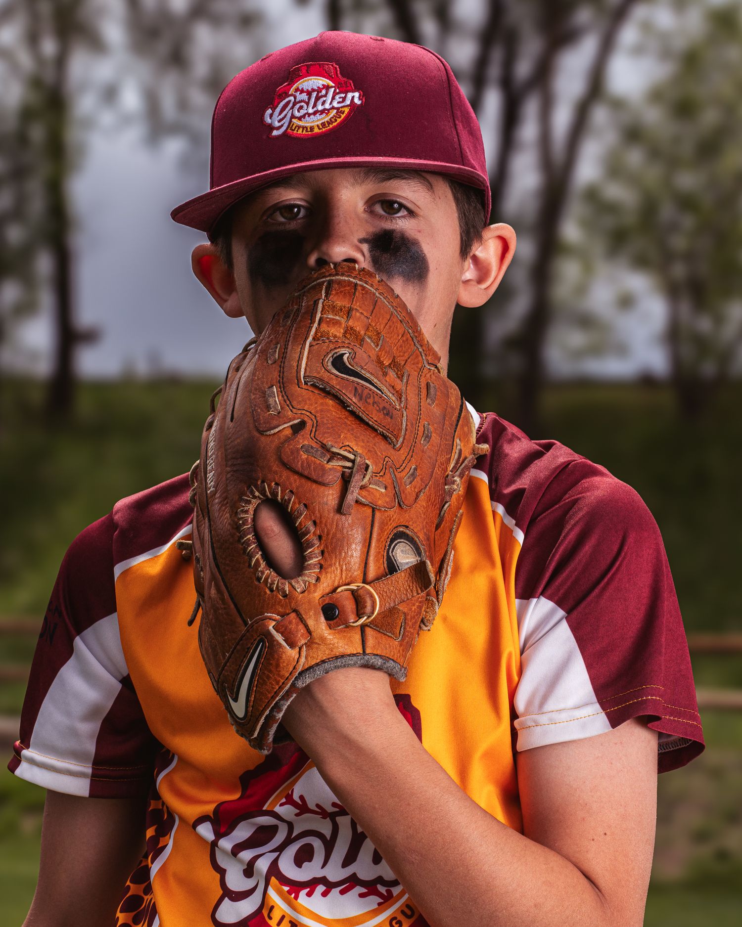 Baseball player wearing burgundy hat and orange jersey peeking out from behind their leather glove during practice.