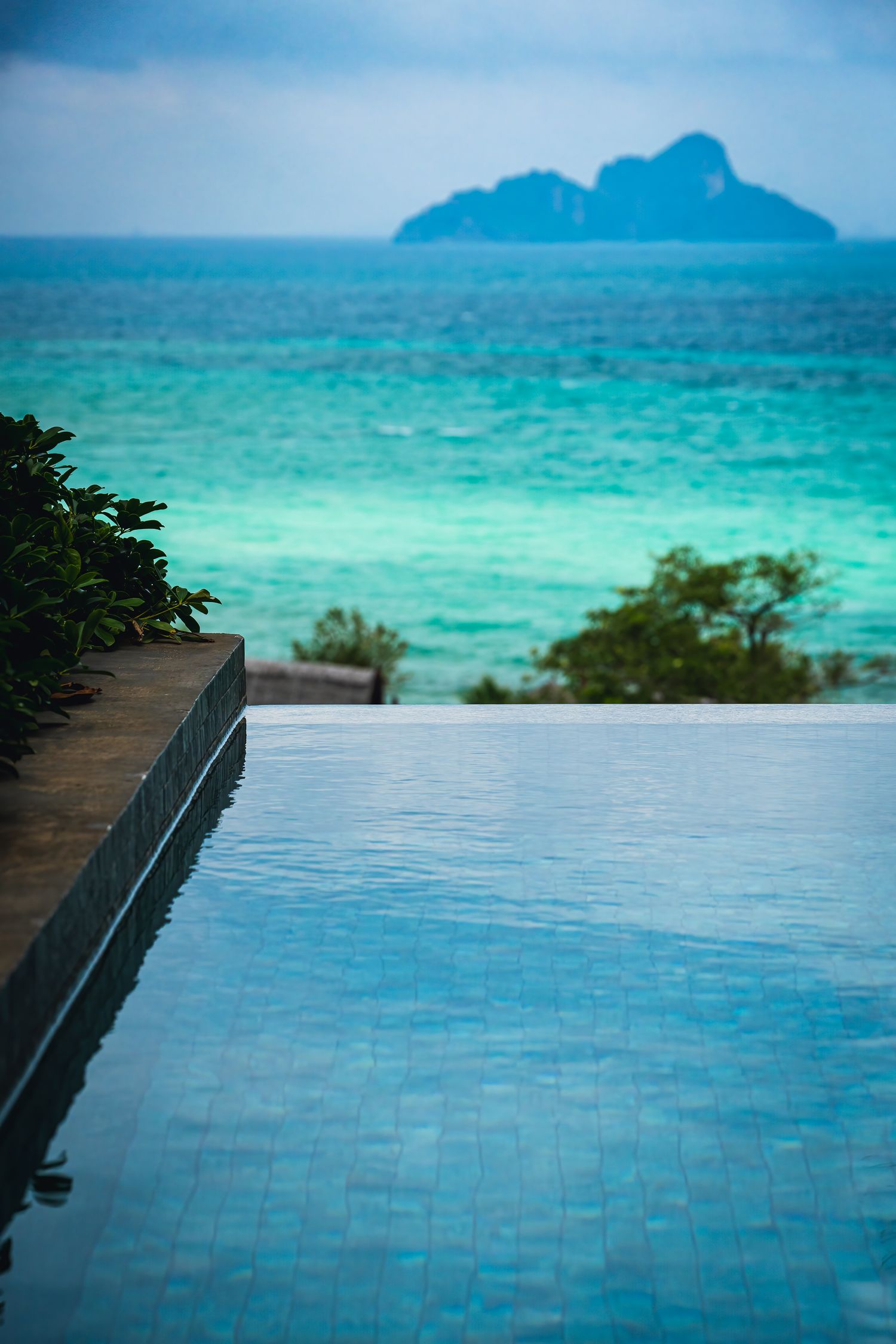 Infinity pool overlooking turquoise tropical ocean with lush greenery and distant island under cloudy sky.