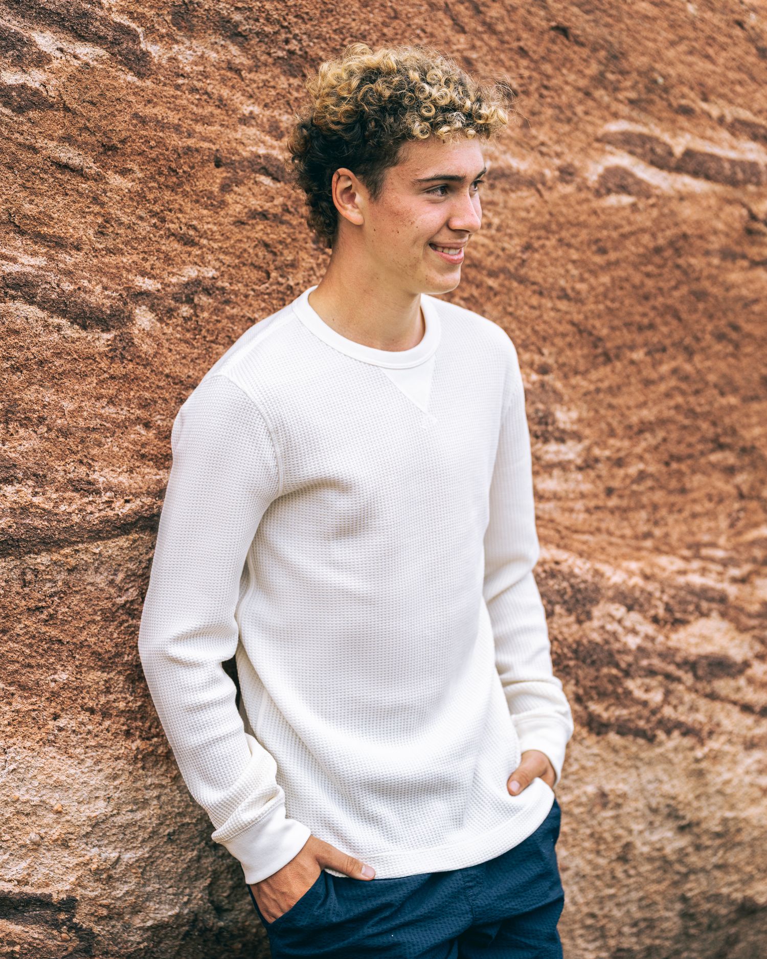 Young man with curly hair wearing a white crew neck sweatshirt, leaning against a rustic red stone wall.