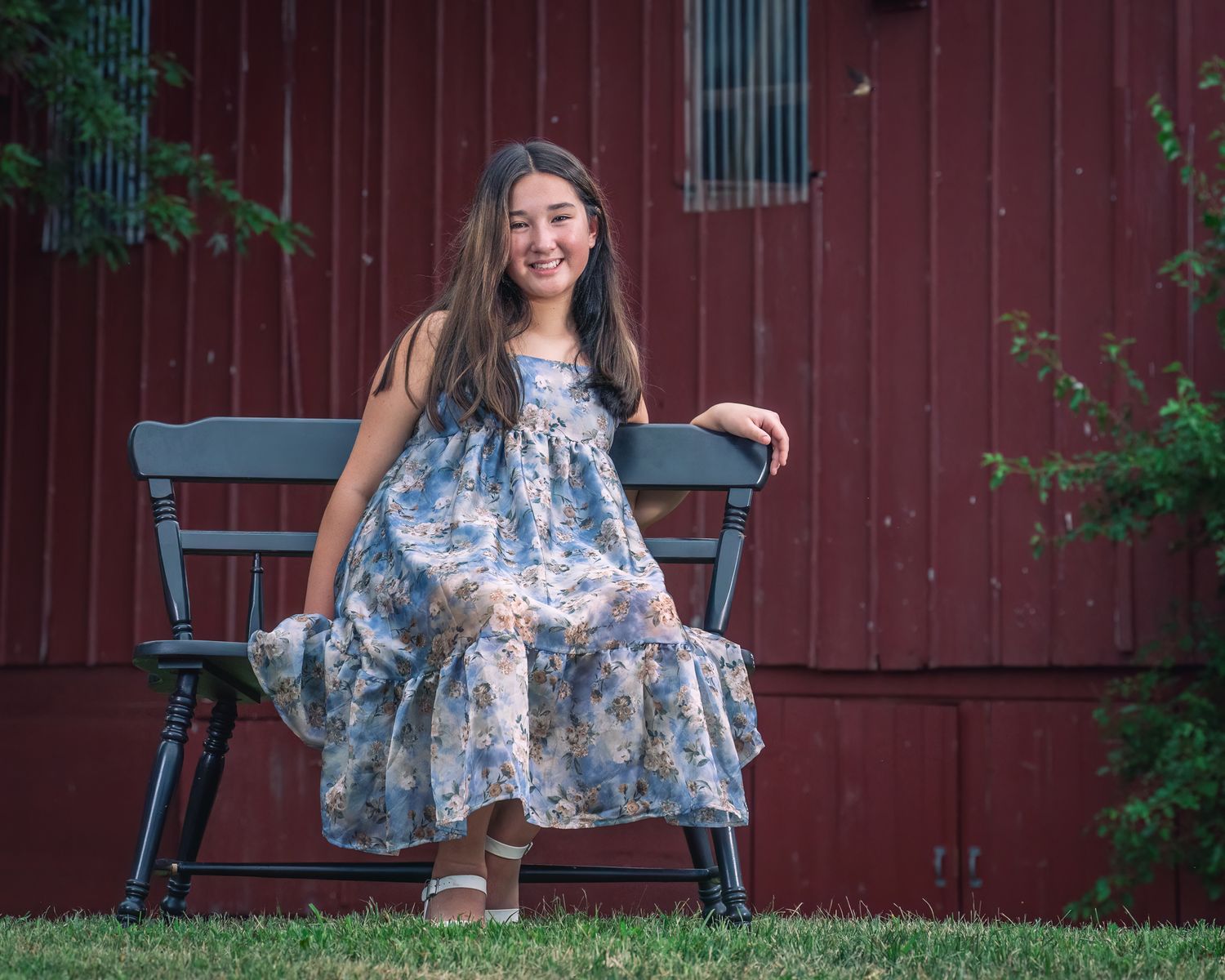 A person in a long blue floral dress poses for a series of outdoor photos in front of a red barn wall.