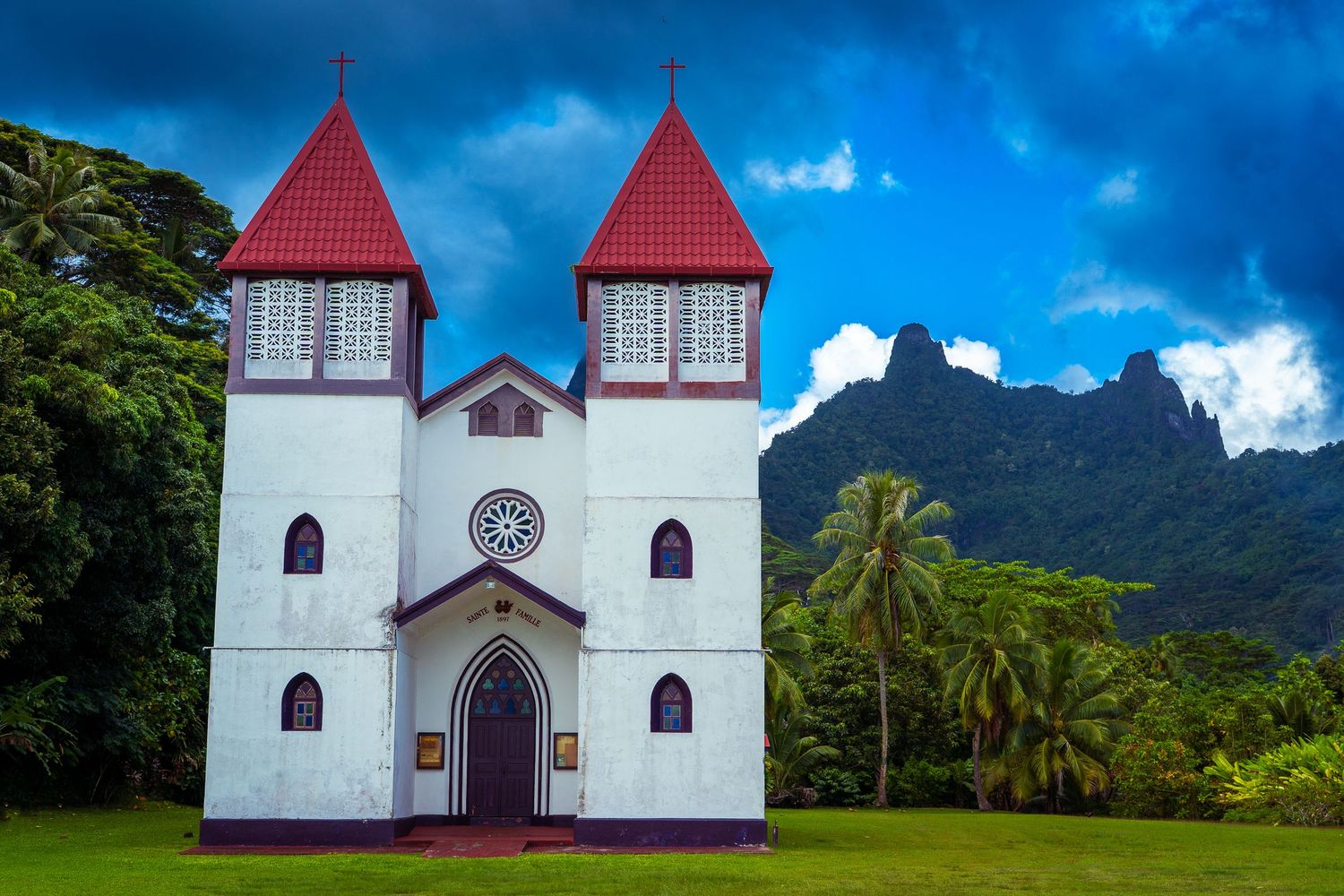 White church with red twin towers set against lush tropical greenery and dramatic mountain backdrop under stormy skies.