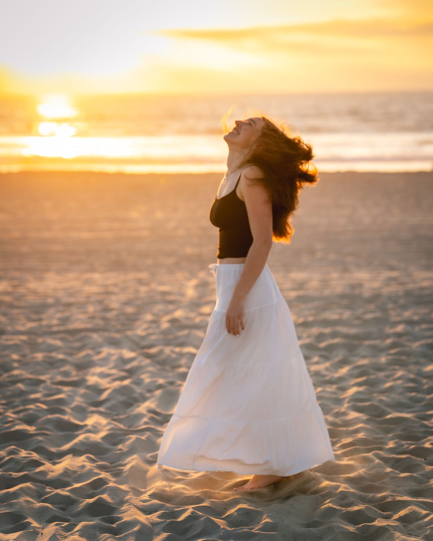 Woman in black top and white maxi skirt standing on beach at golden sunset with hair blowing in wind.