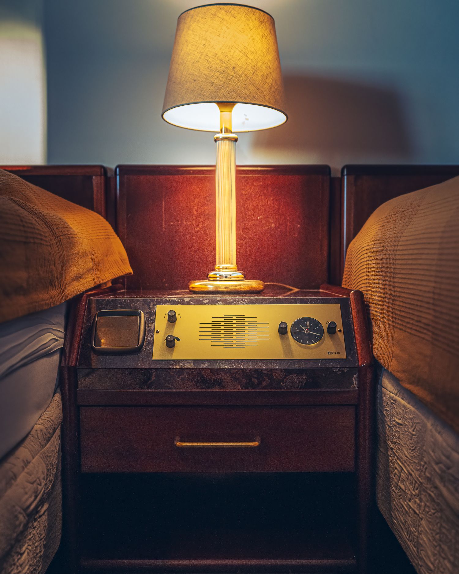 Vintage motel nightstand with retro yellow radio, analog clock, small TV, and glowing table lamp between two beds.