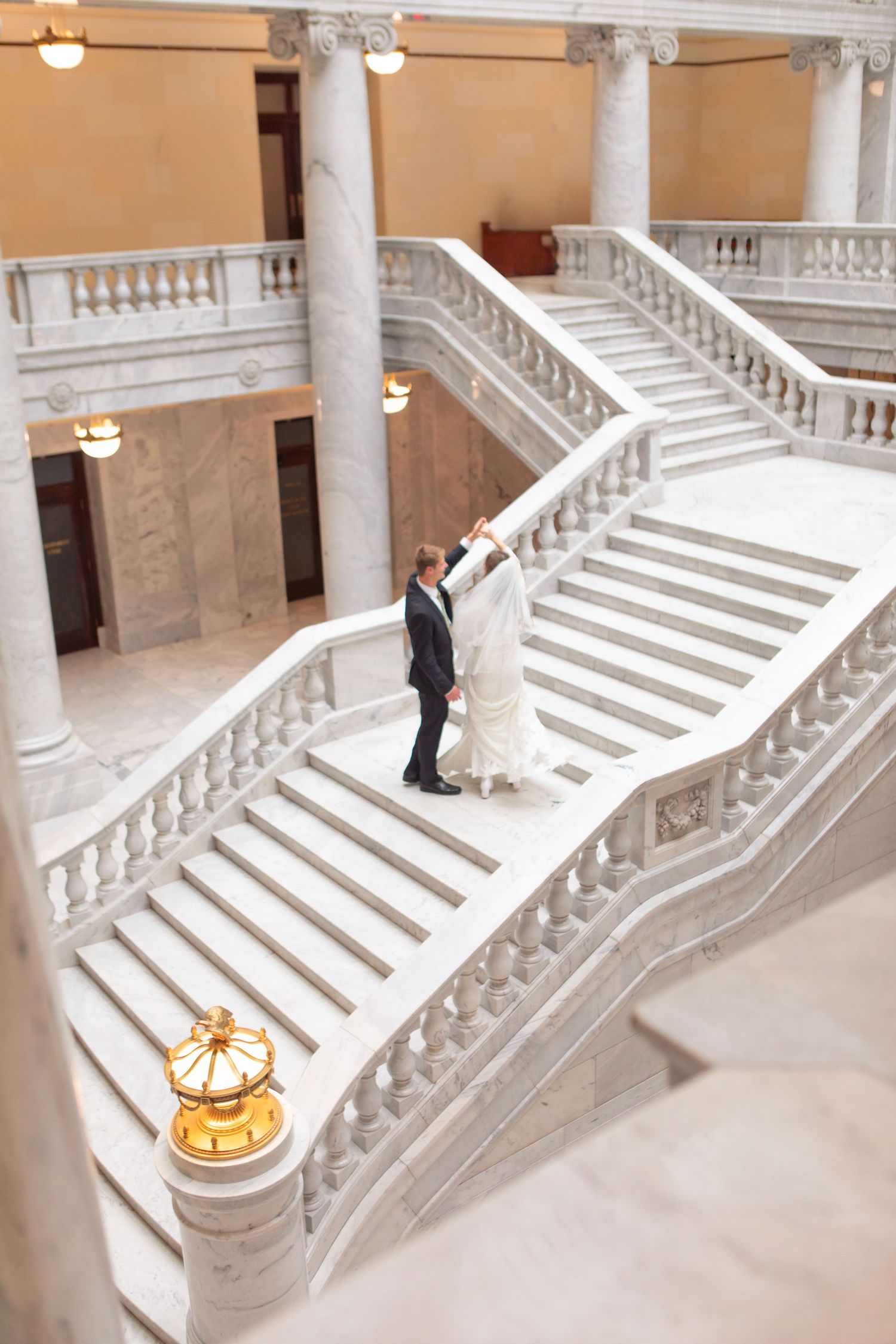 Bride and groom dancing on grand marble staircase in elegant white stone building with classical columns.