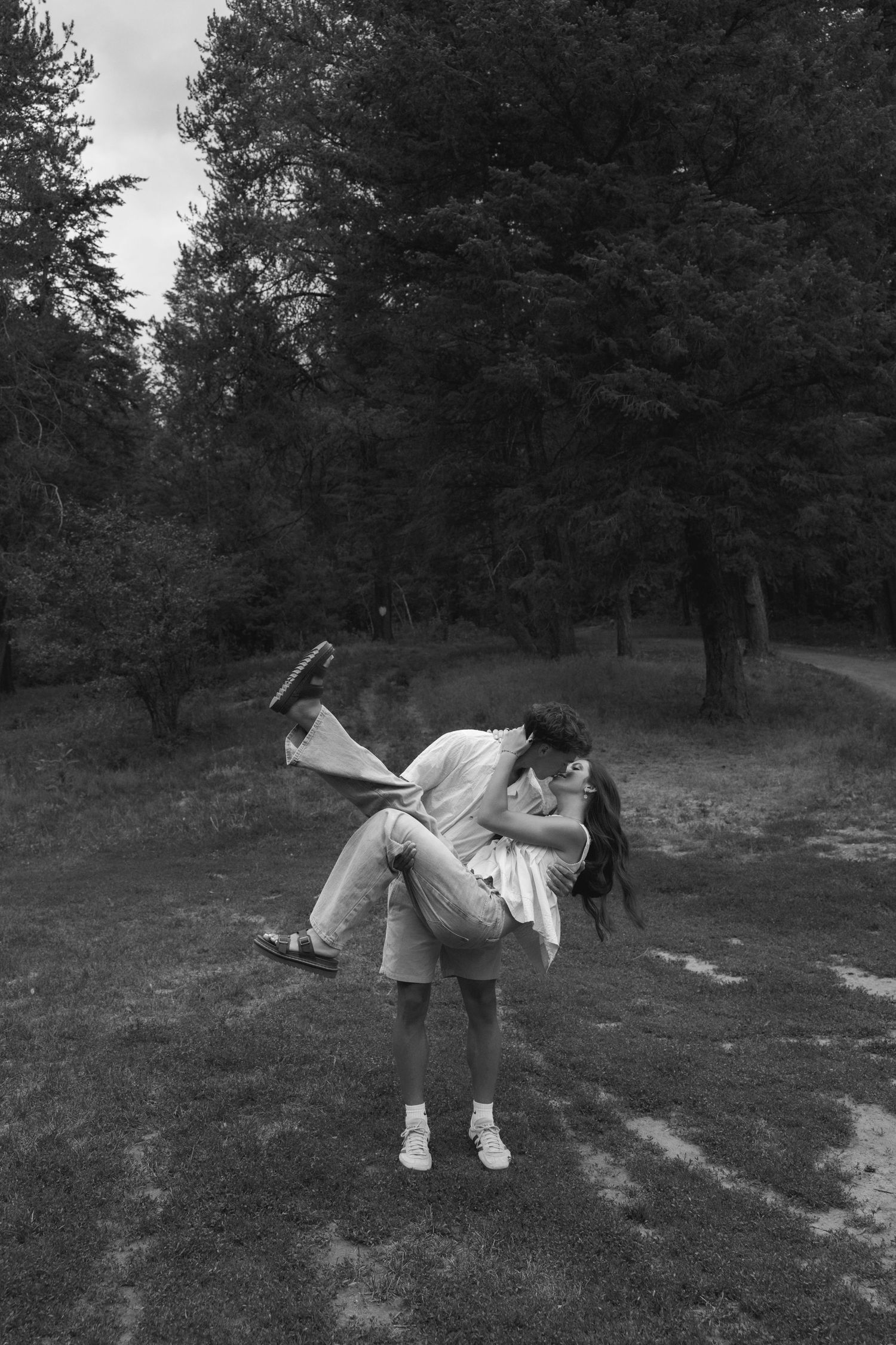 Black and white photo of a couple kissing outdoors, man lifting woman in a romantic embrace surrounded by lush trees.
