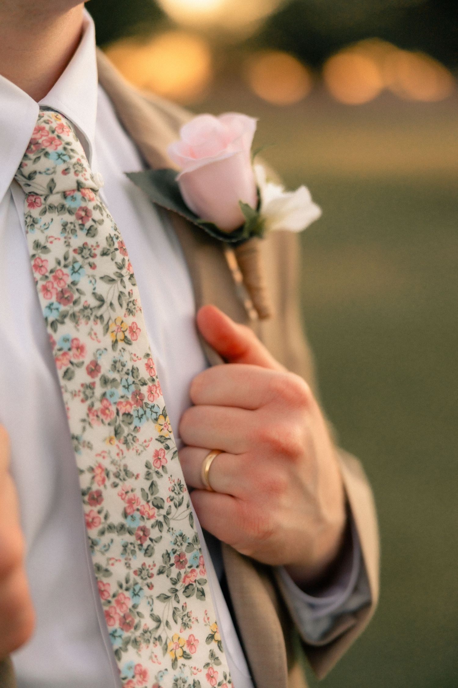 Groom in tan suit with floral tie and pink rose boutonniere, wearing a gold wedding band.