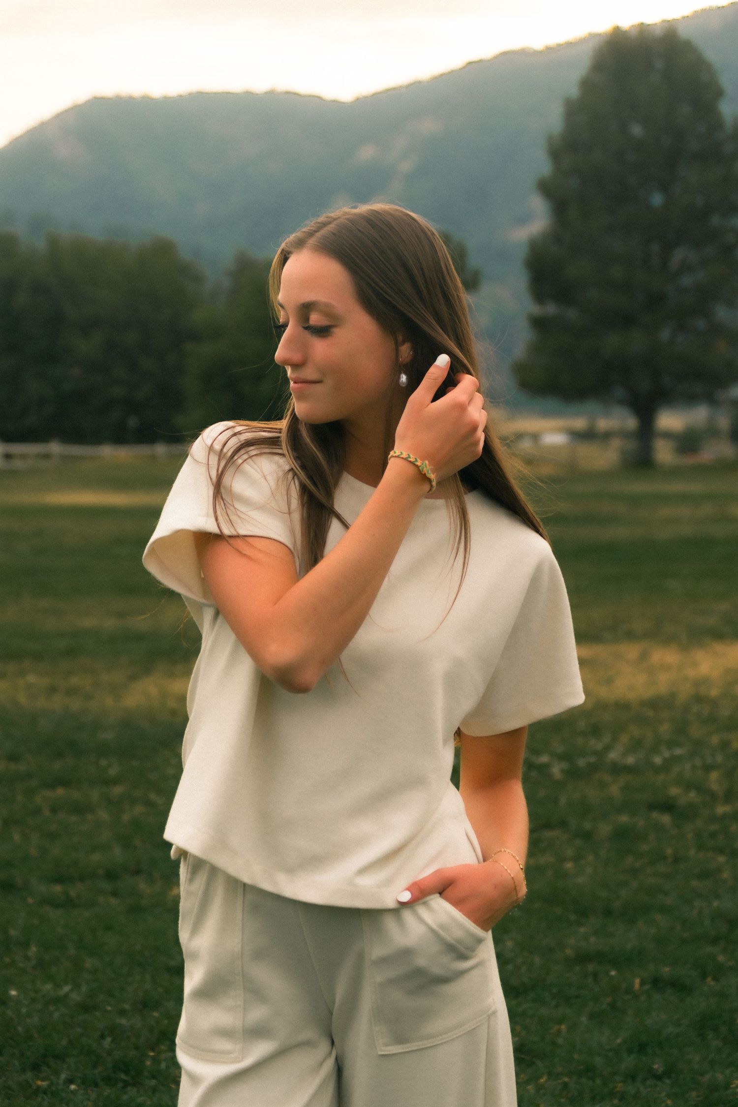 Young woman in cream outfit poses outdoors with mountain backdrop, touching her hair gracefully.