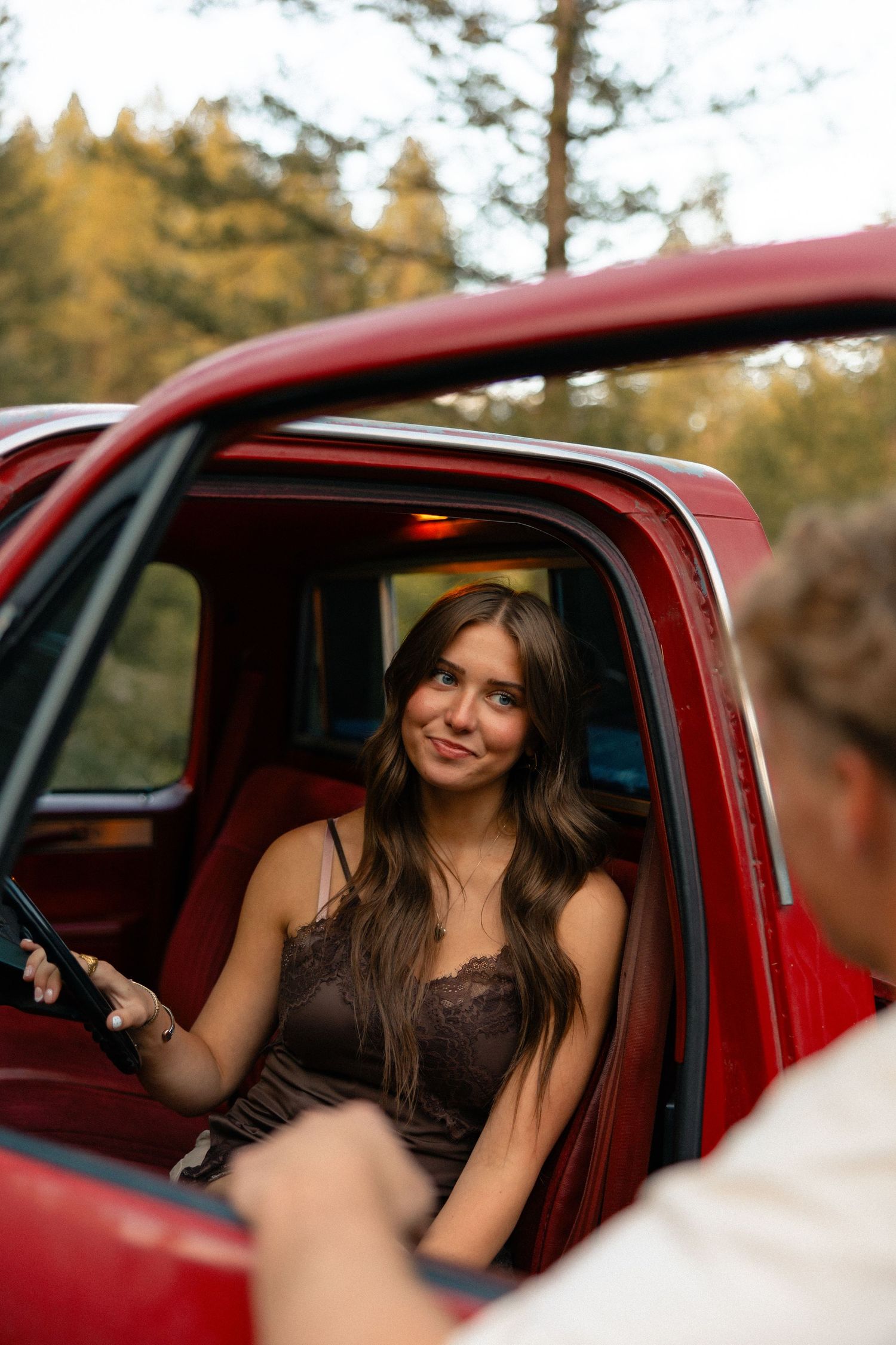Young woman with long brown hair in a brown lace top smiling from a classic red pickup truck in a forest setting.