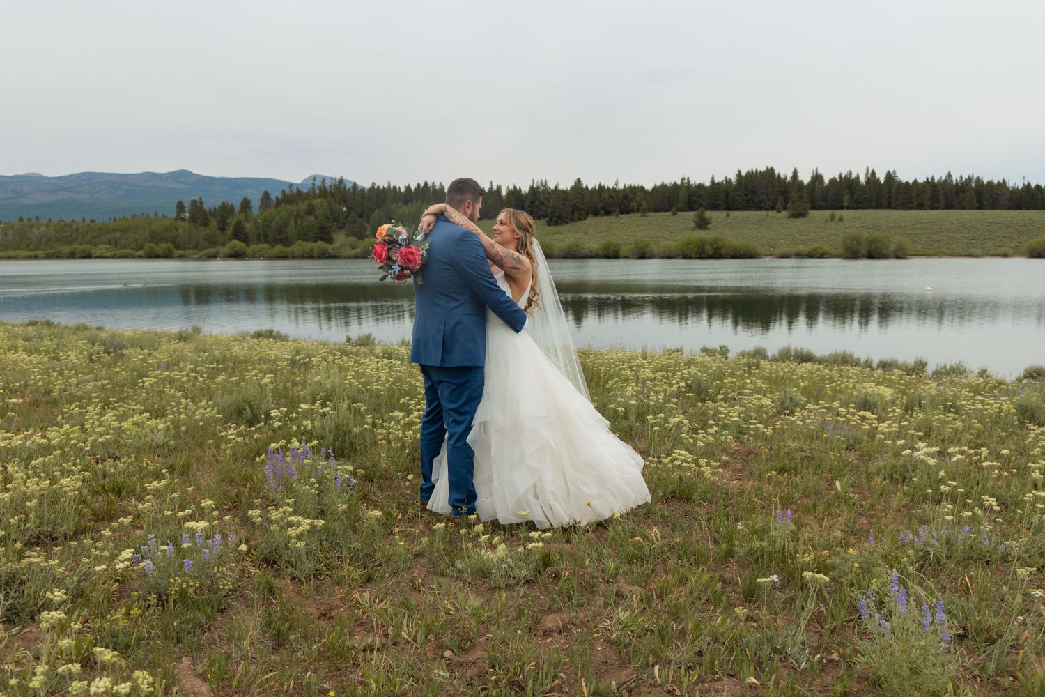 A couple in wedding attire stands in a field of wildflowers near a lake with mountains in the background.