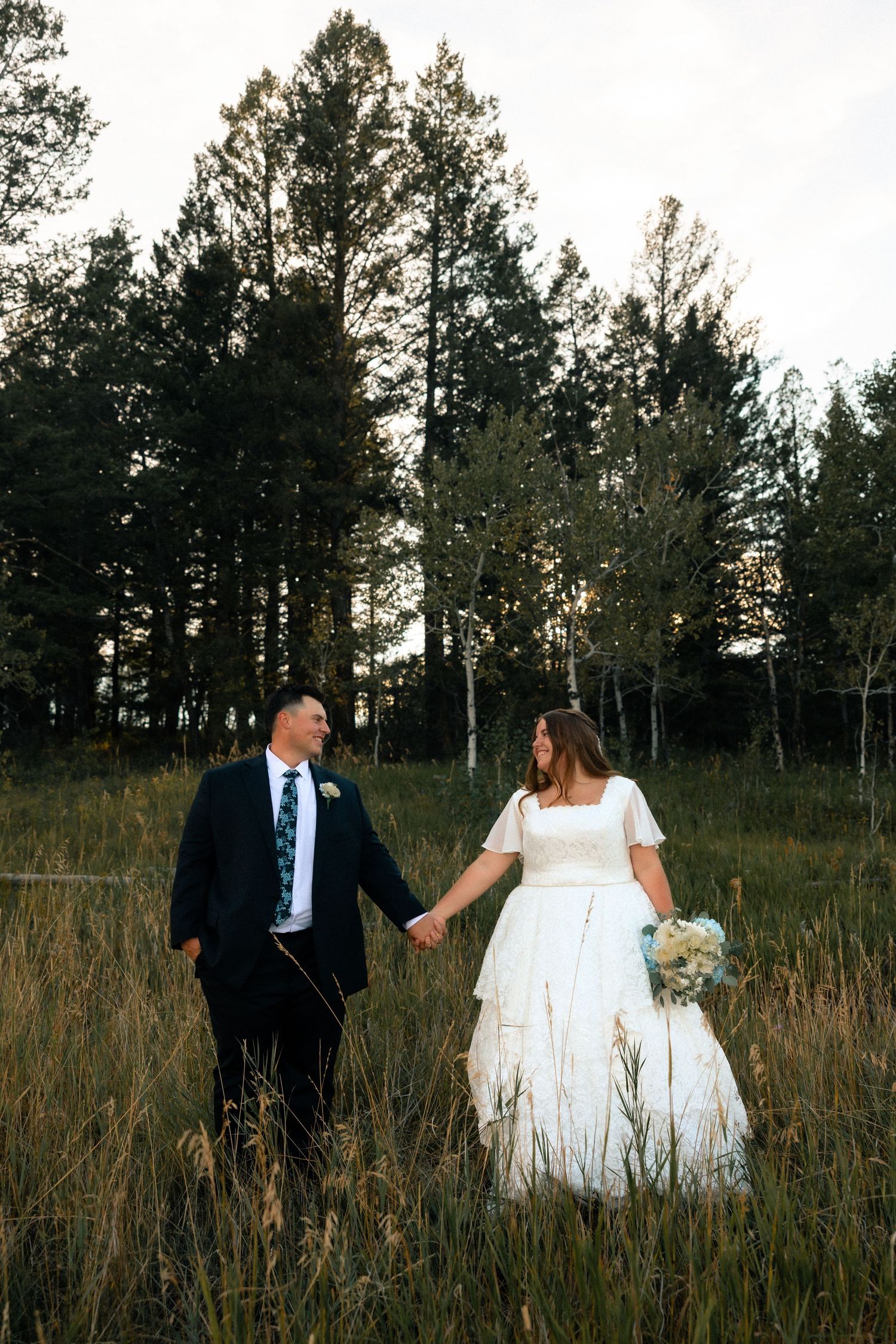 Bride and groom hold hands walking through a sunlit meadow with tall pine trees in the background.