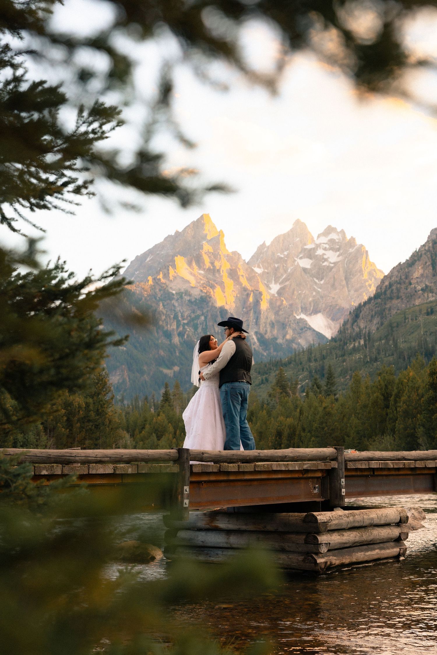 Couple embracing on a wooden dock at sunset with the Grand Teton mountains and forest in the background.