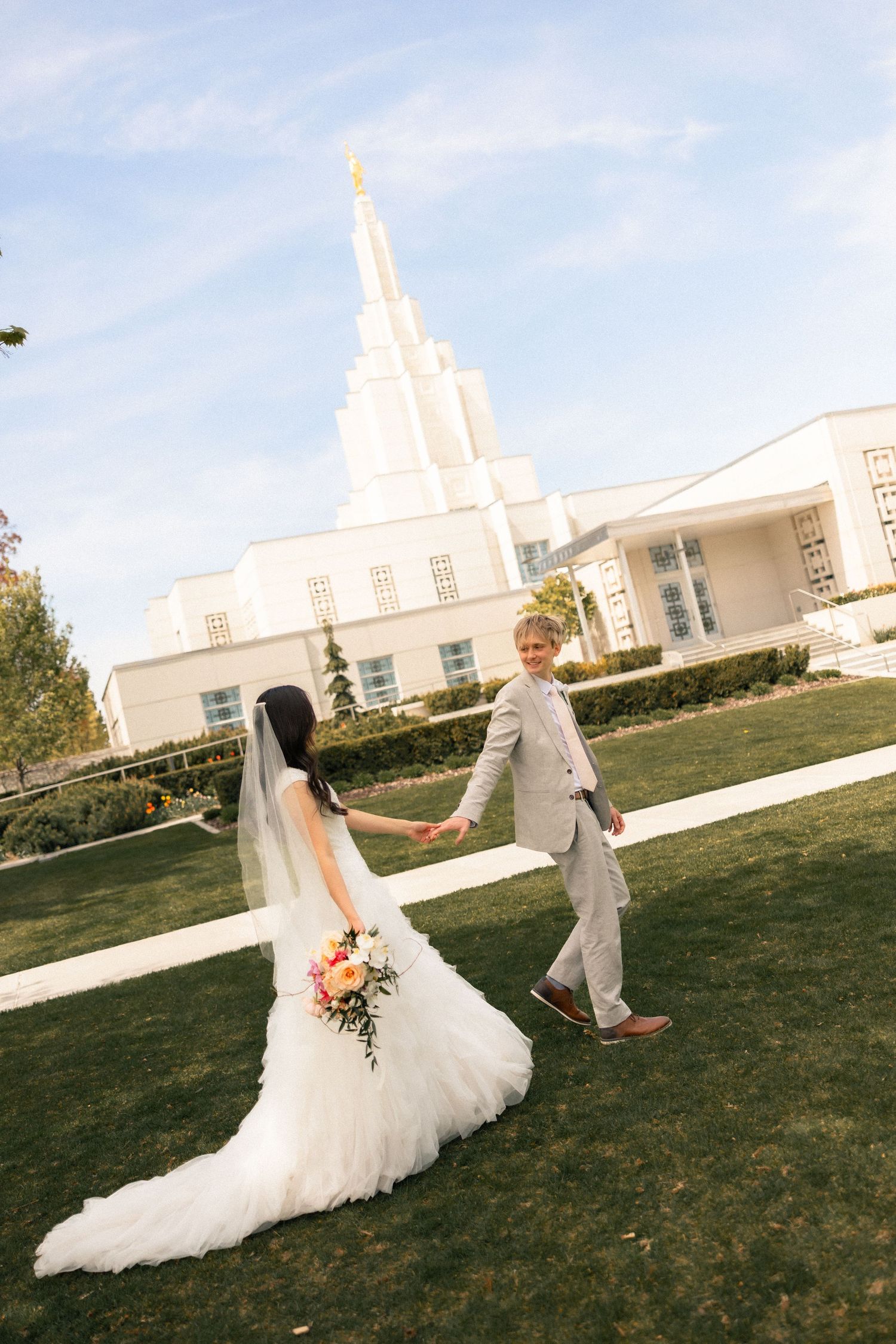 Bride and groom holding hands walking on green lawn outside a white LDS temple on their wedding day.