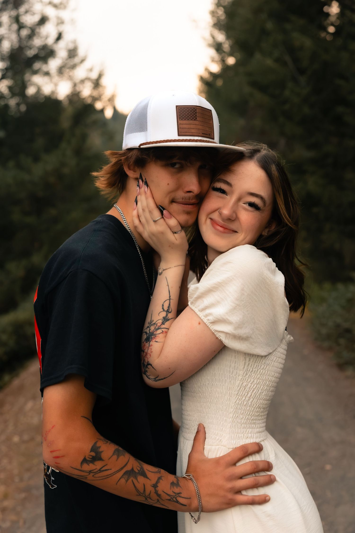 A couple embraces on a forest path, woman in white dress smiling, man with tattoos and trucker hat holding her close.