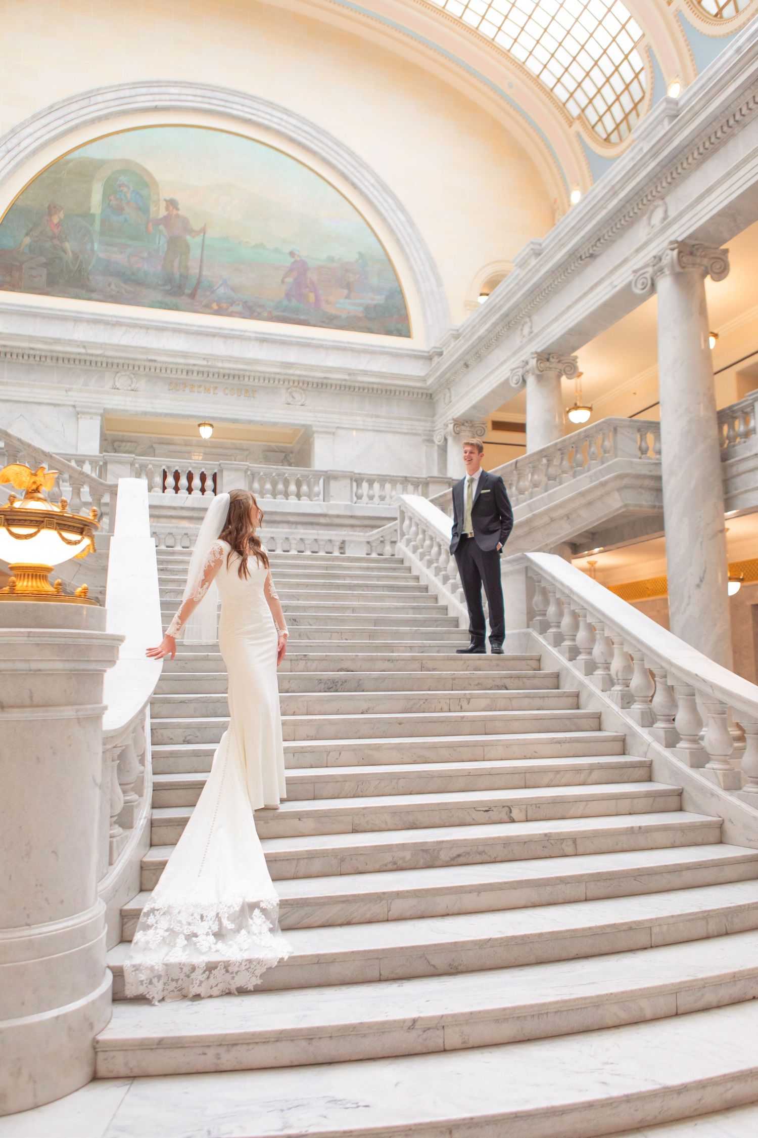Bride and groom on grand marble staircase in elegant building with classical columns and ornate mural.