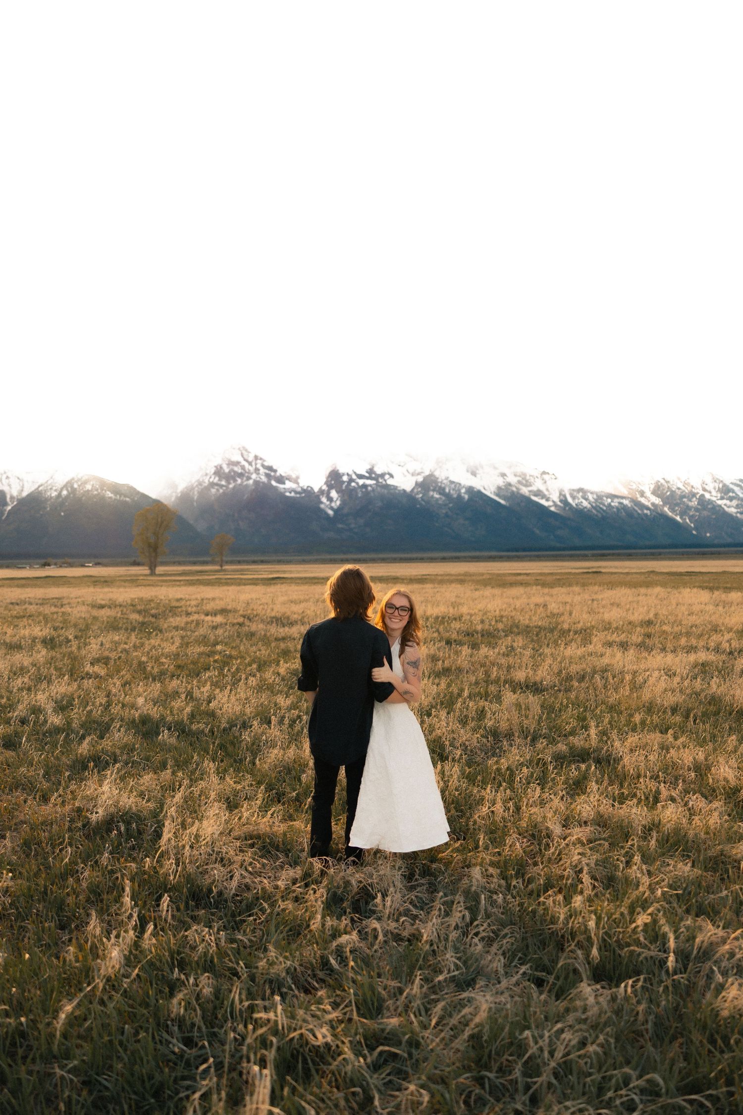 Bride and groom embrace in golden field with snow-capped Grand Teton mountains at sunset.