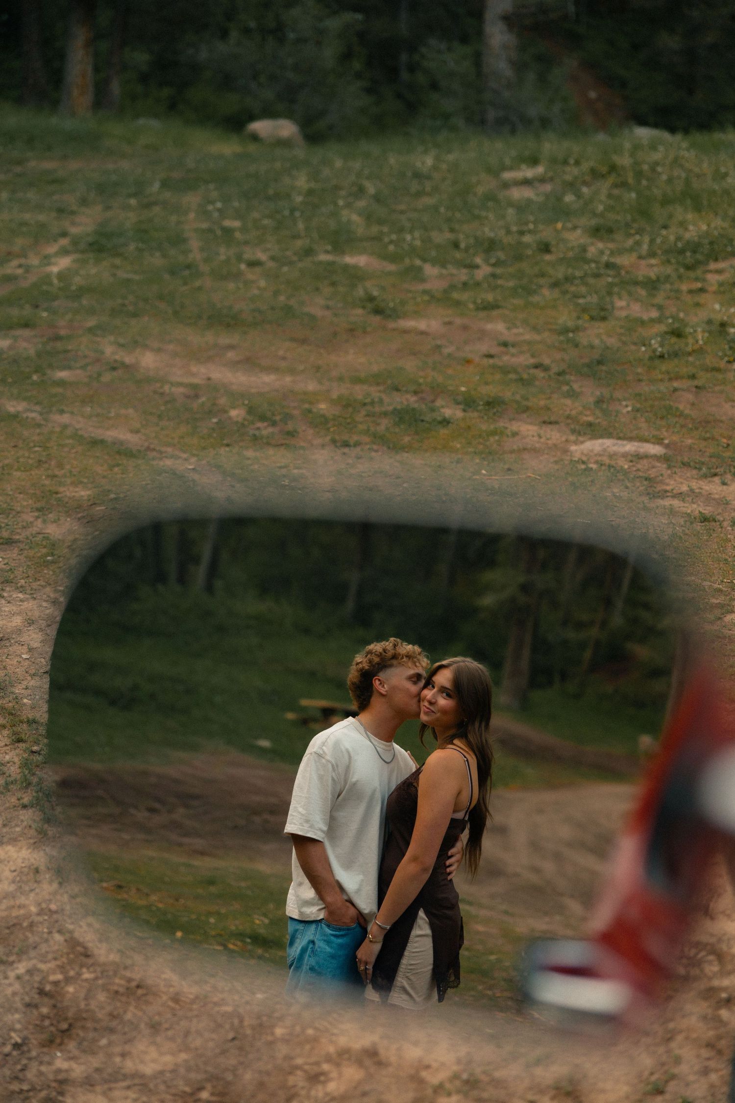 Couple holding hands and kissing reflected in a car side mirror with a green forest background.