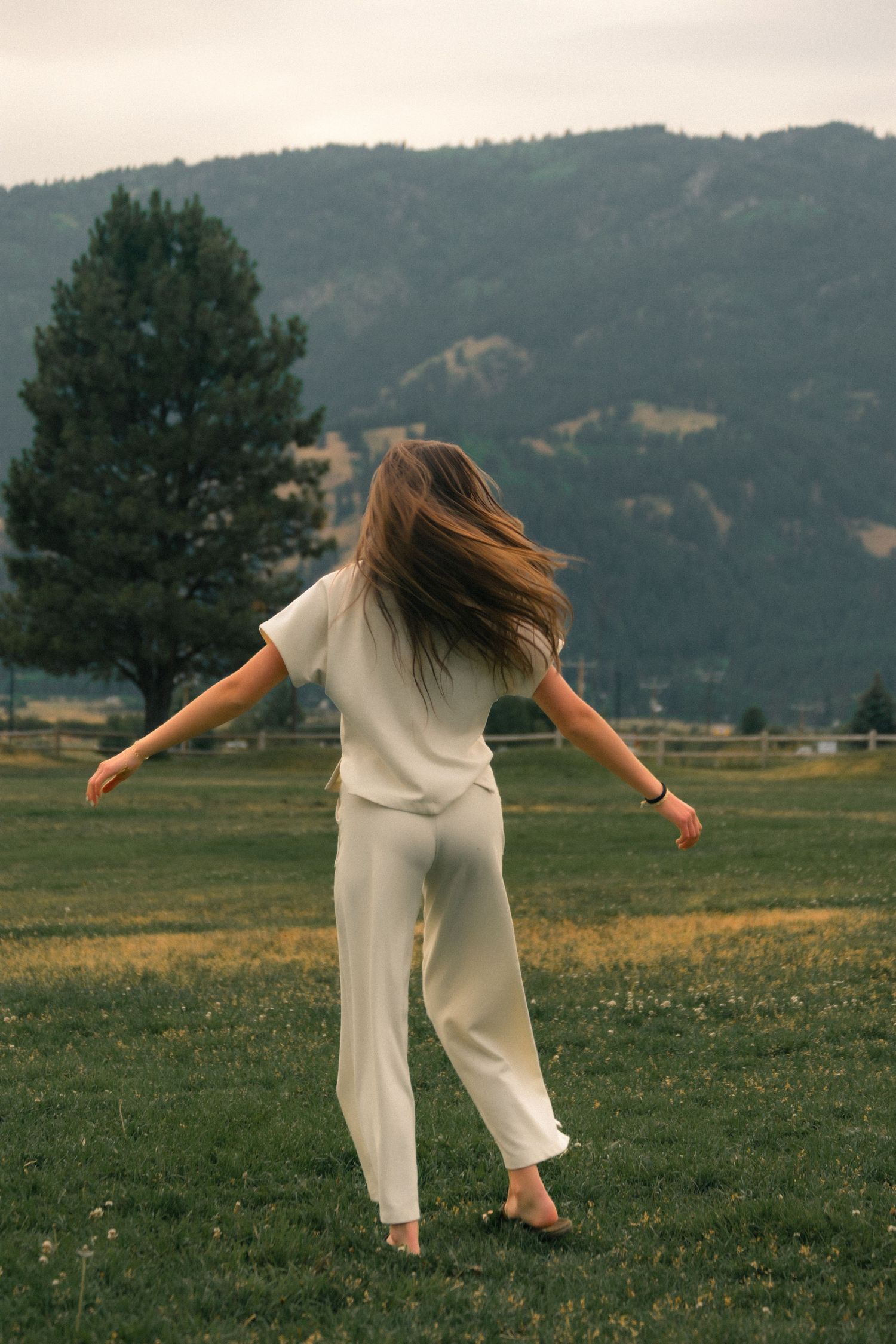 Woman in white outfit standing in green field with arms outstretched, hair blowing in wind, mountains in background.