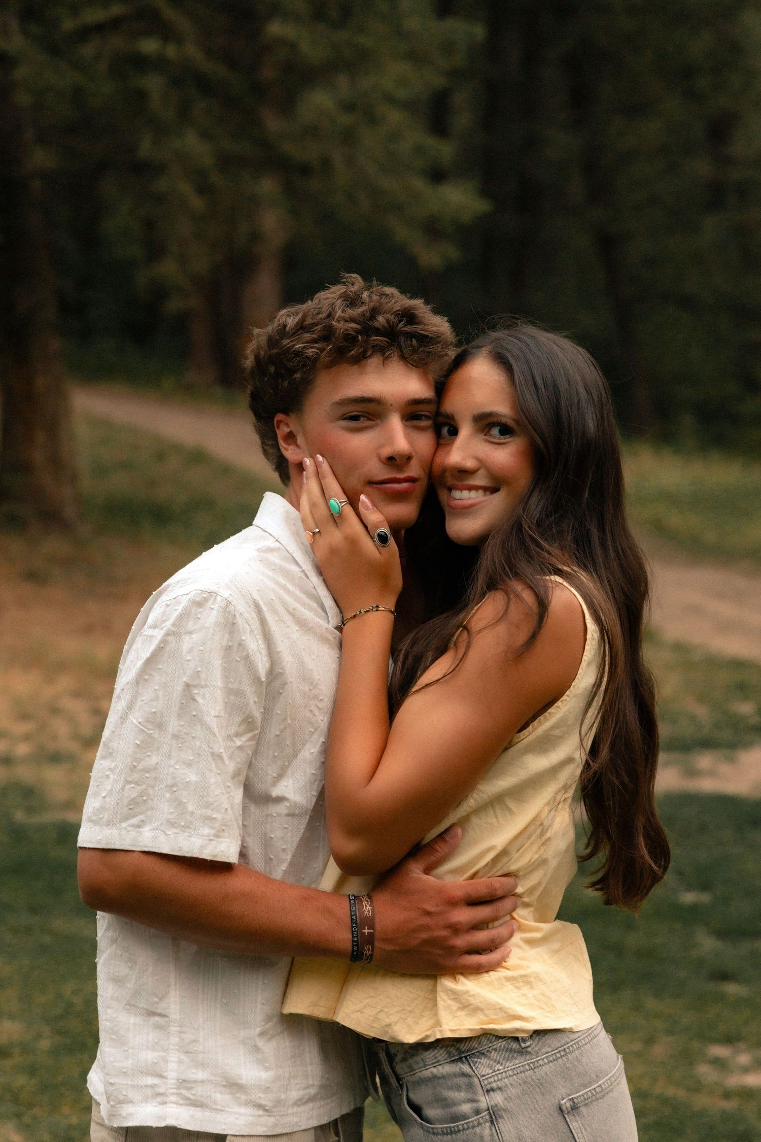 A young couple embracing outdoors, smiling warmly, with lush green trees in the background.