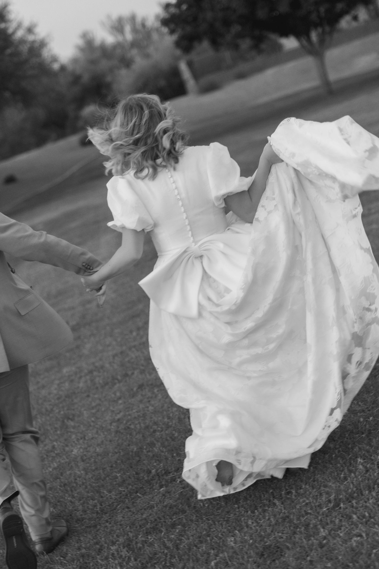 Black and white photo of a bride in a flowing white gown holding hands with groom while walking outdoors.
