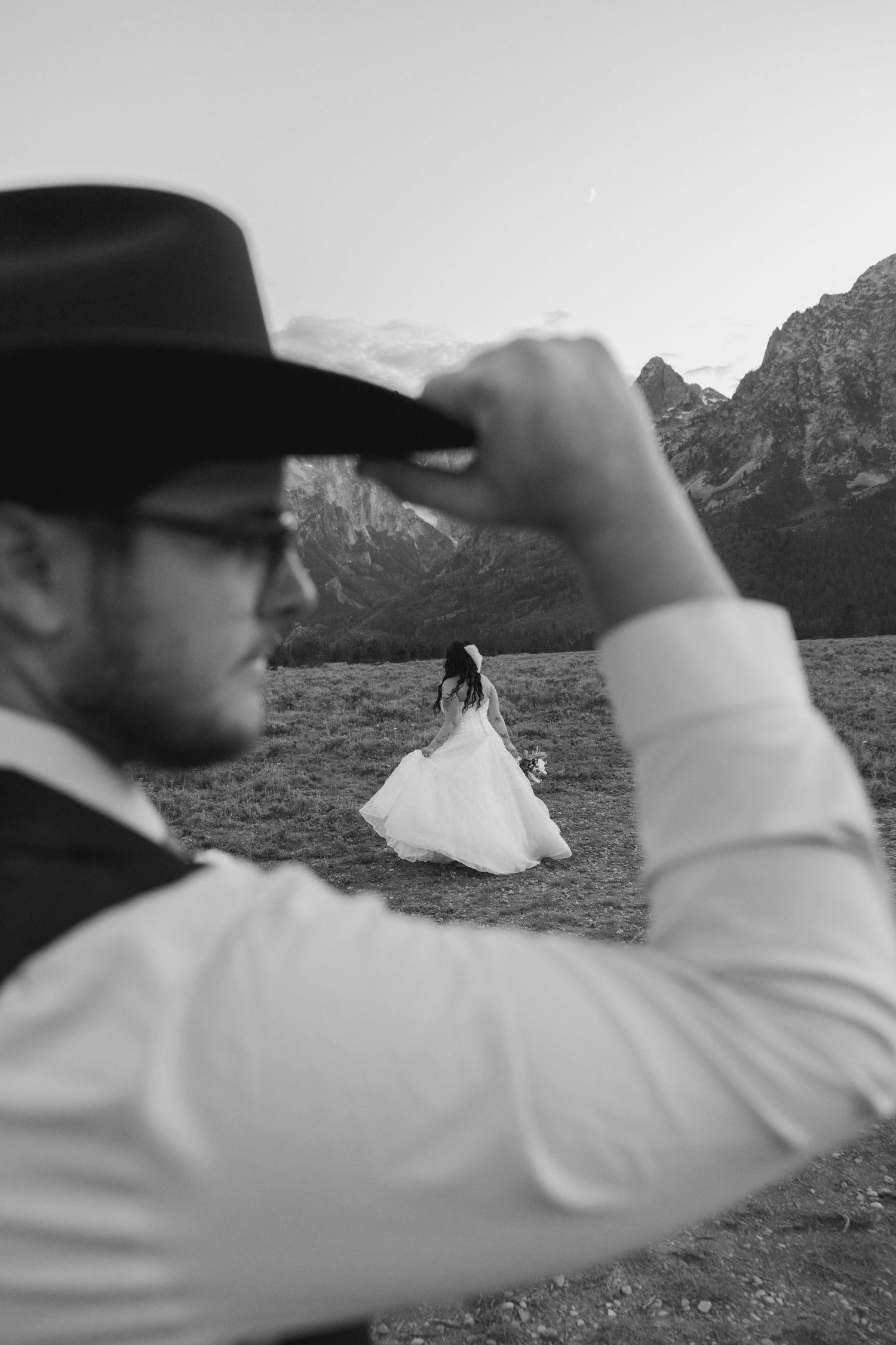 Black and white photo of groom adjusting cowboy hat with bride in white dress standing in mountain field.