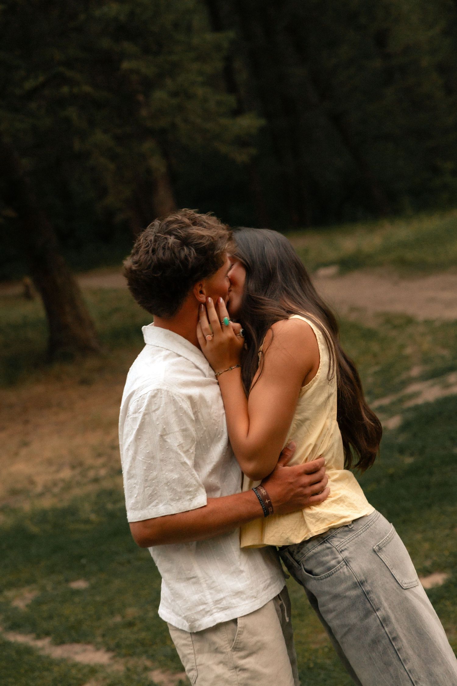 Couple sharing a romantic kiss in a lush green park, woman in yellow top and jeans tenderly holding mans face.