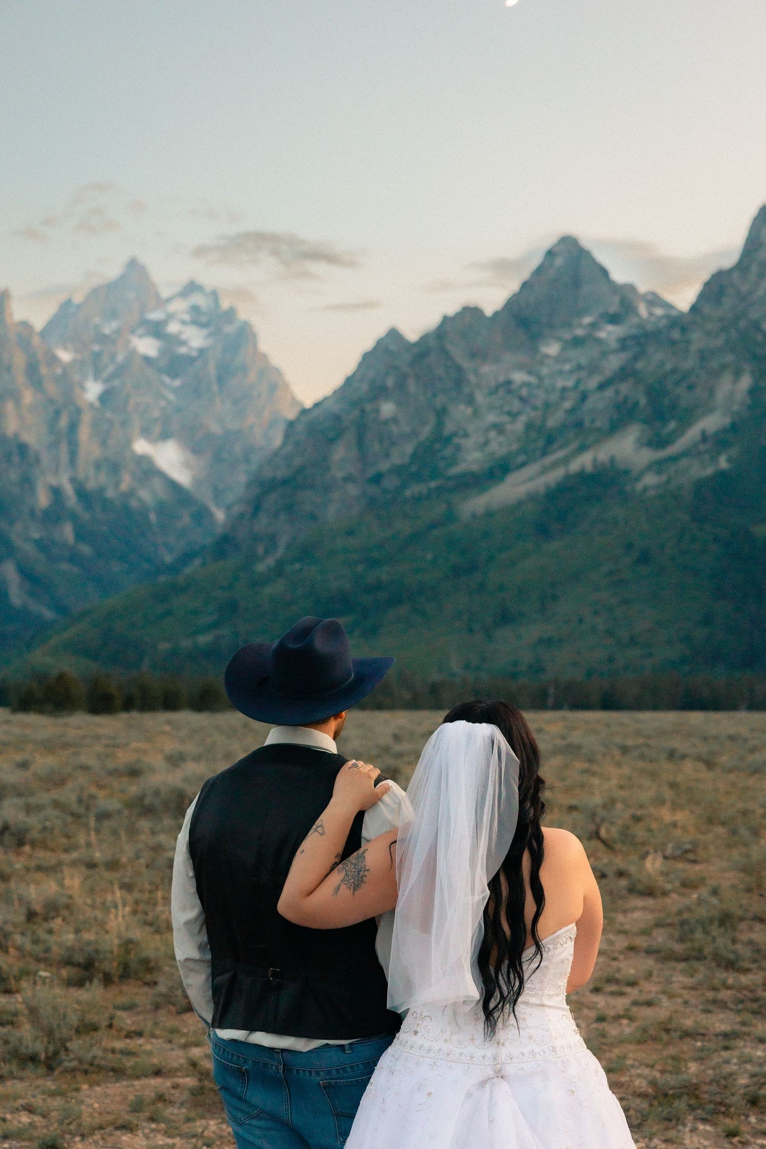 A bride and groom stand arm in arm facing the Grand Teton mountains at sunset during their wedding day.