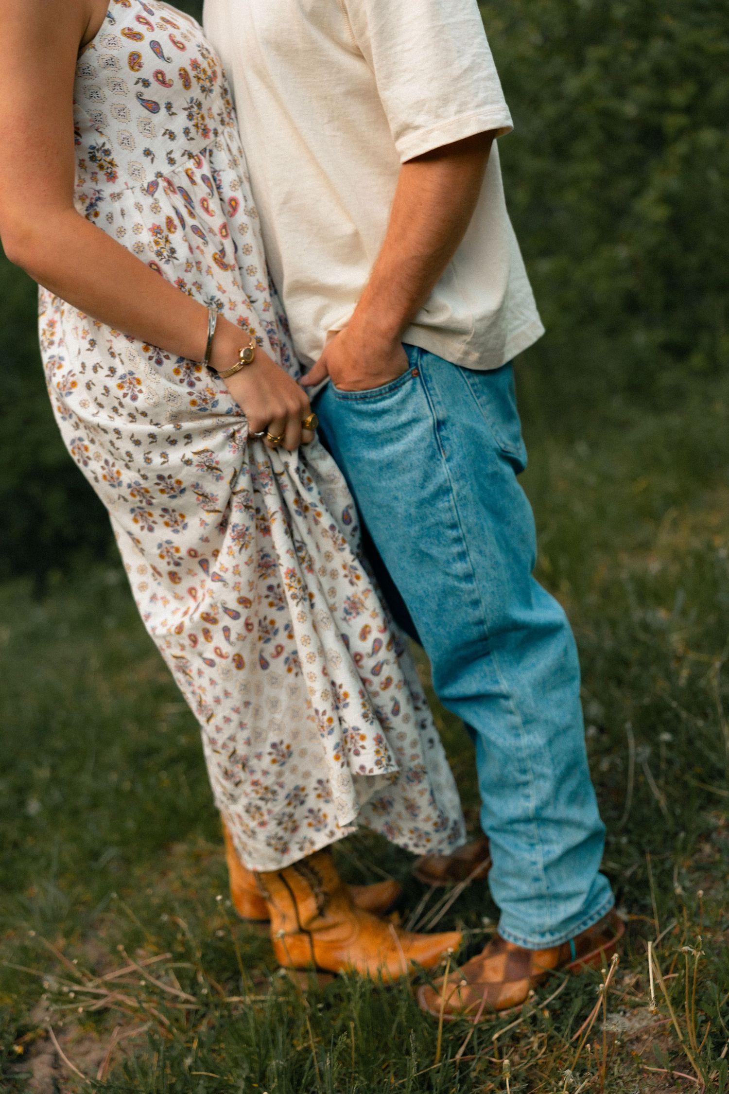 Close-up of a couple holding hands outdoors, woman in floral dress and cowboy boots, man in jeans and white t-shirt.
