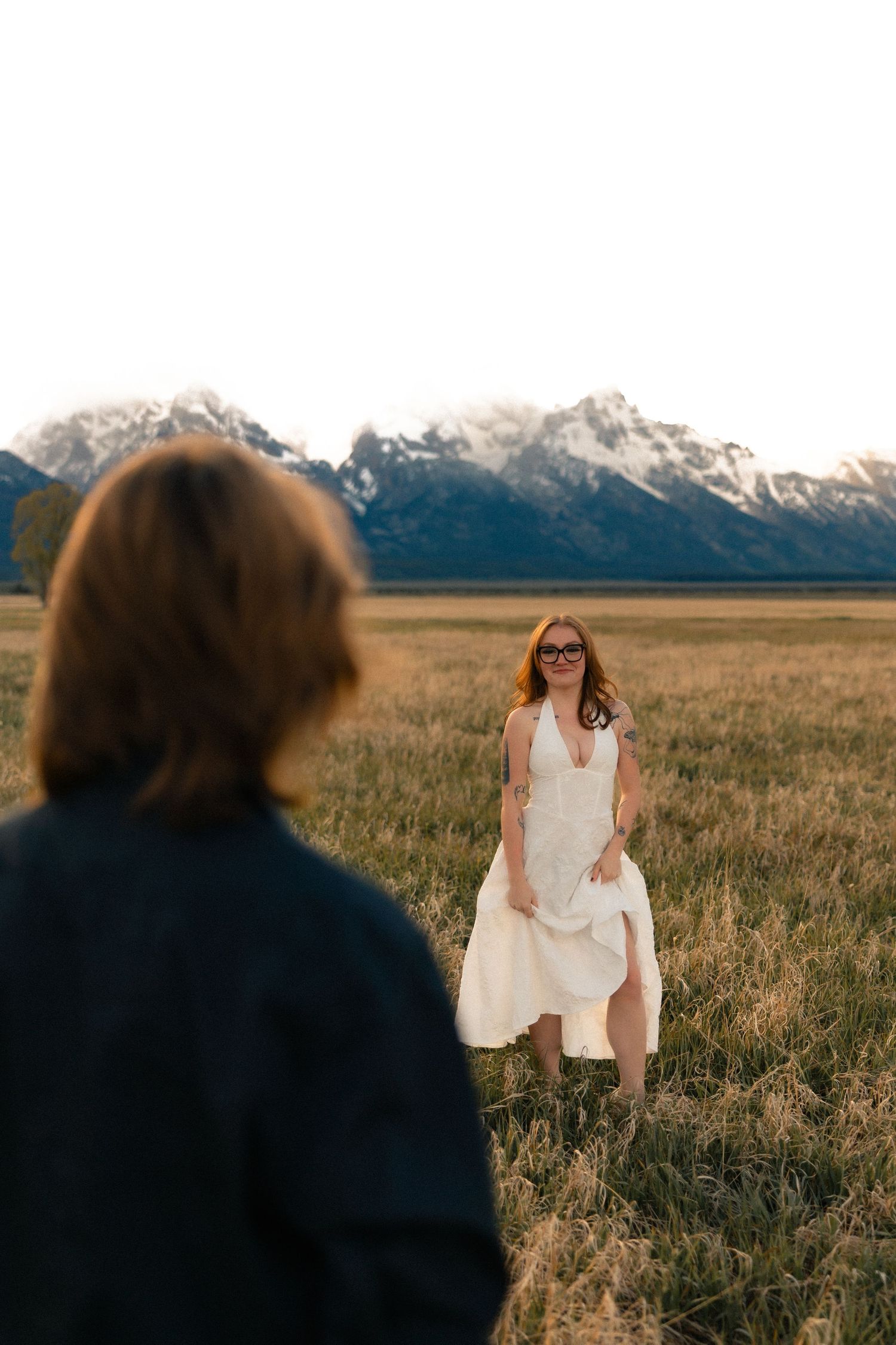 Bride in white halter dress standing in mountain meadow with groom in foreground, Grand Tetons backdrop.