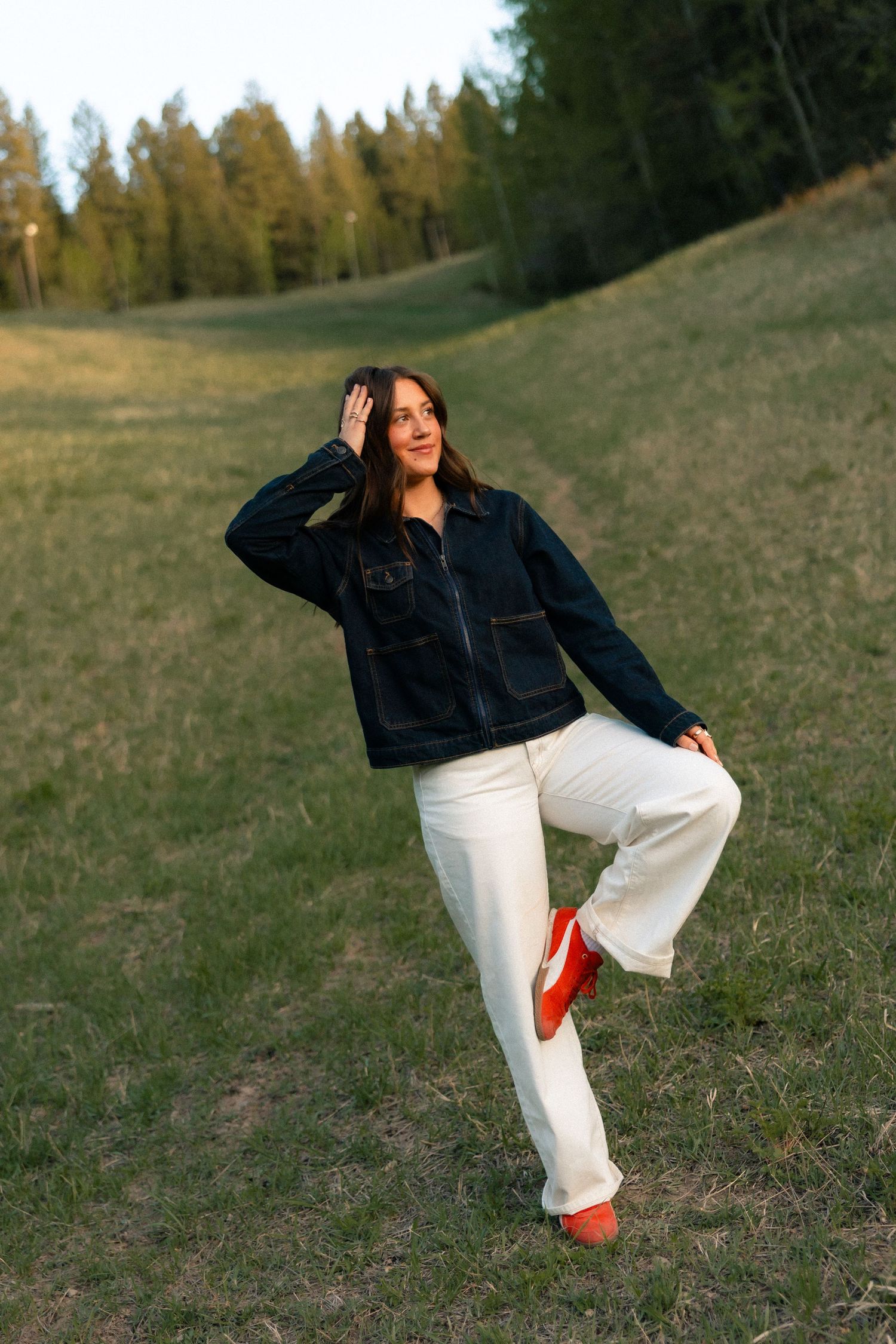 Woman in dark denim jacket, white pants, and red shoes poses playfully on a grassy hillside with forest backdrop.