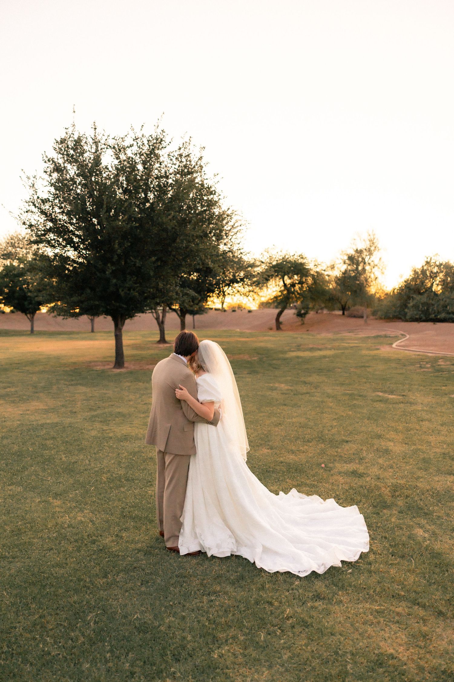 Bride and groom embrace at sunset on lush green lawn, her white gown trailing behind them near trees.