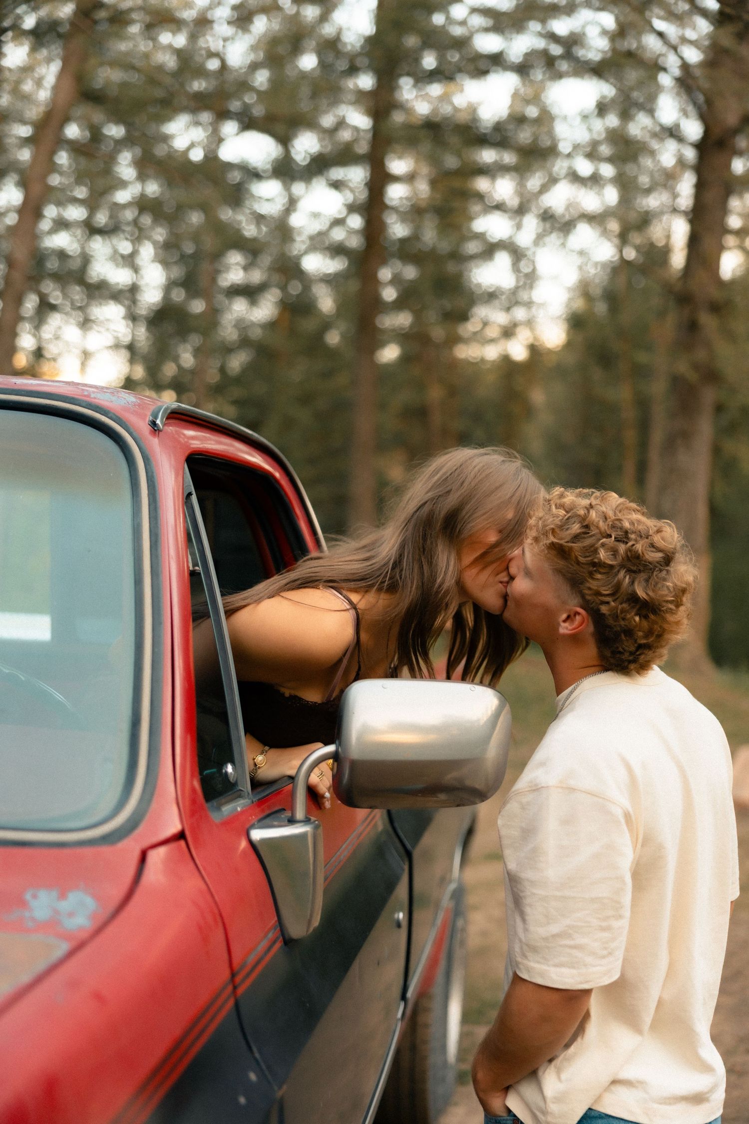 Couple sharing a romantic kiss through a red truck window surrounded by tall pine trees in a forest setting.