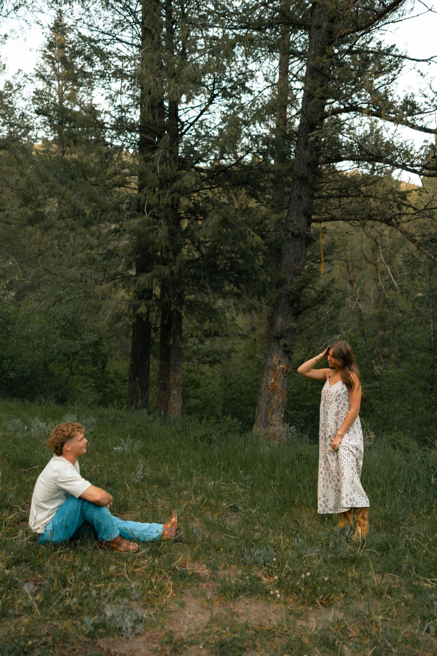 A young couple in a forest meadow, man sitting cross-legged on grass, woman standing in floral dress among tall pine trees.