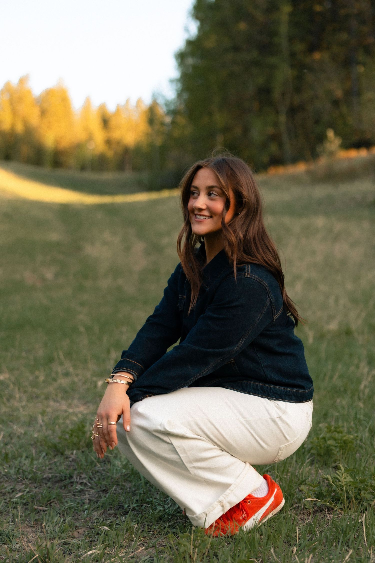 Smiling young woman in black jacket and white pants crouching on grass field with autumn trees in background.