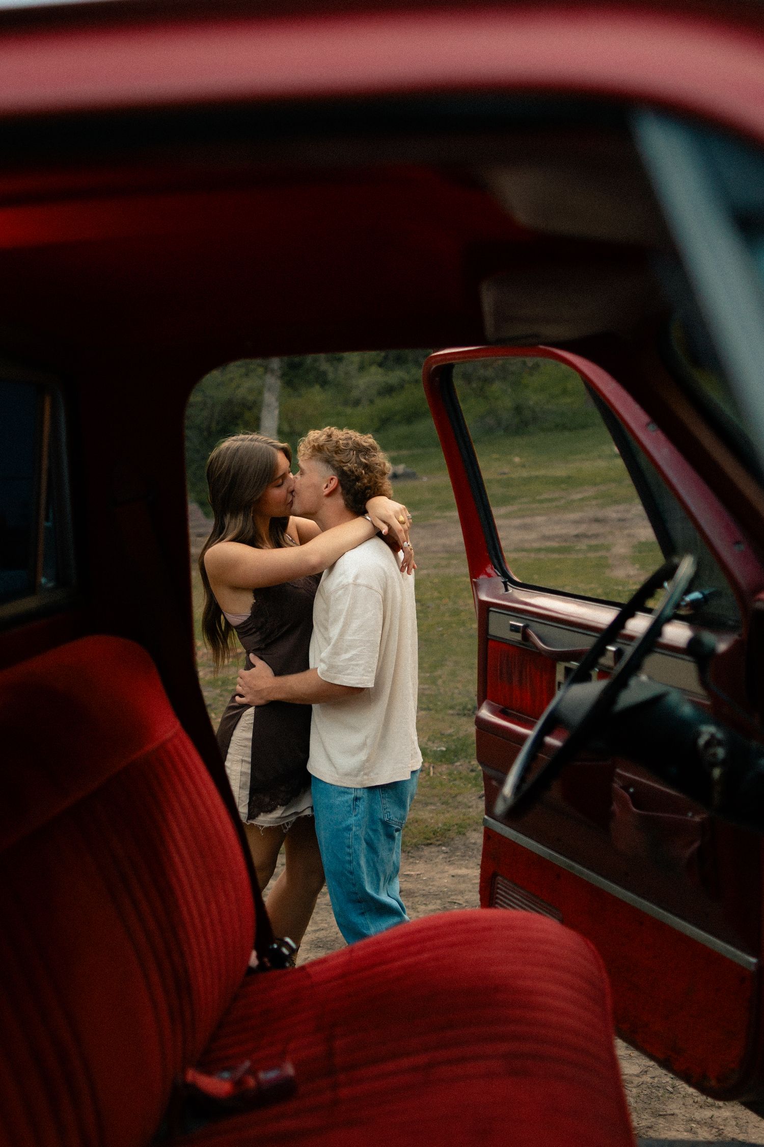 A couple shares a romantic kiss outside a vintage red truck, framed by the open door and red interior seats.
