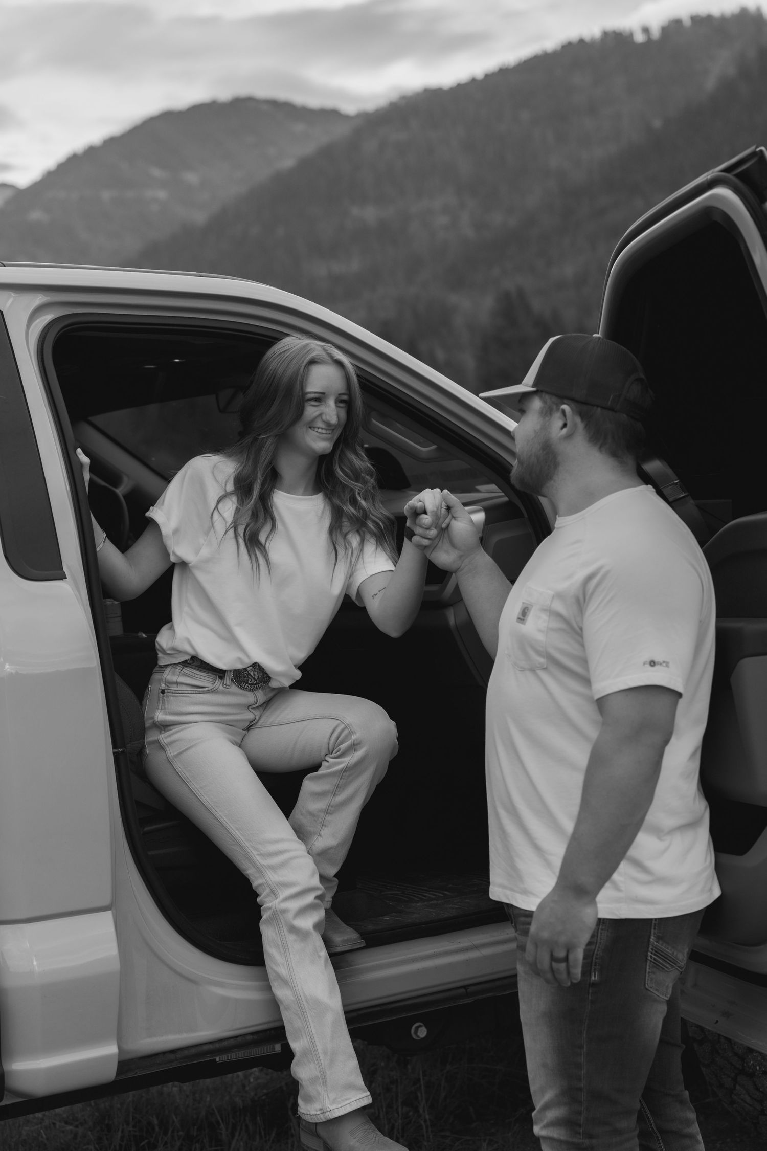 Black and white photo of a smiling couple sharing a moment by an open car door with mountains in the background.