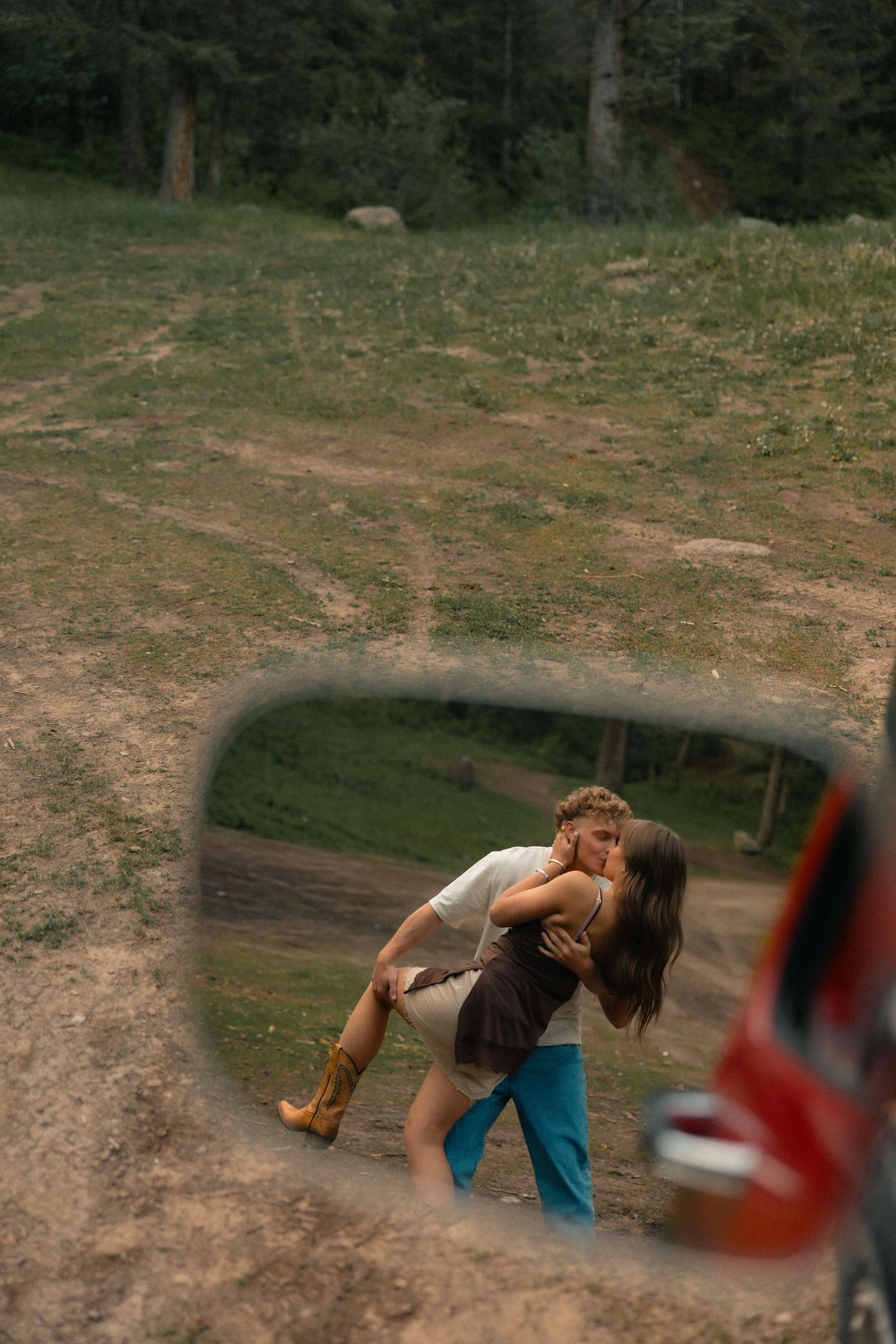 Couple sharing a romantic kiss reflected in a car side mirror, surrounded by a lush green forest landscape.