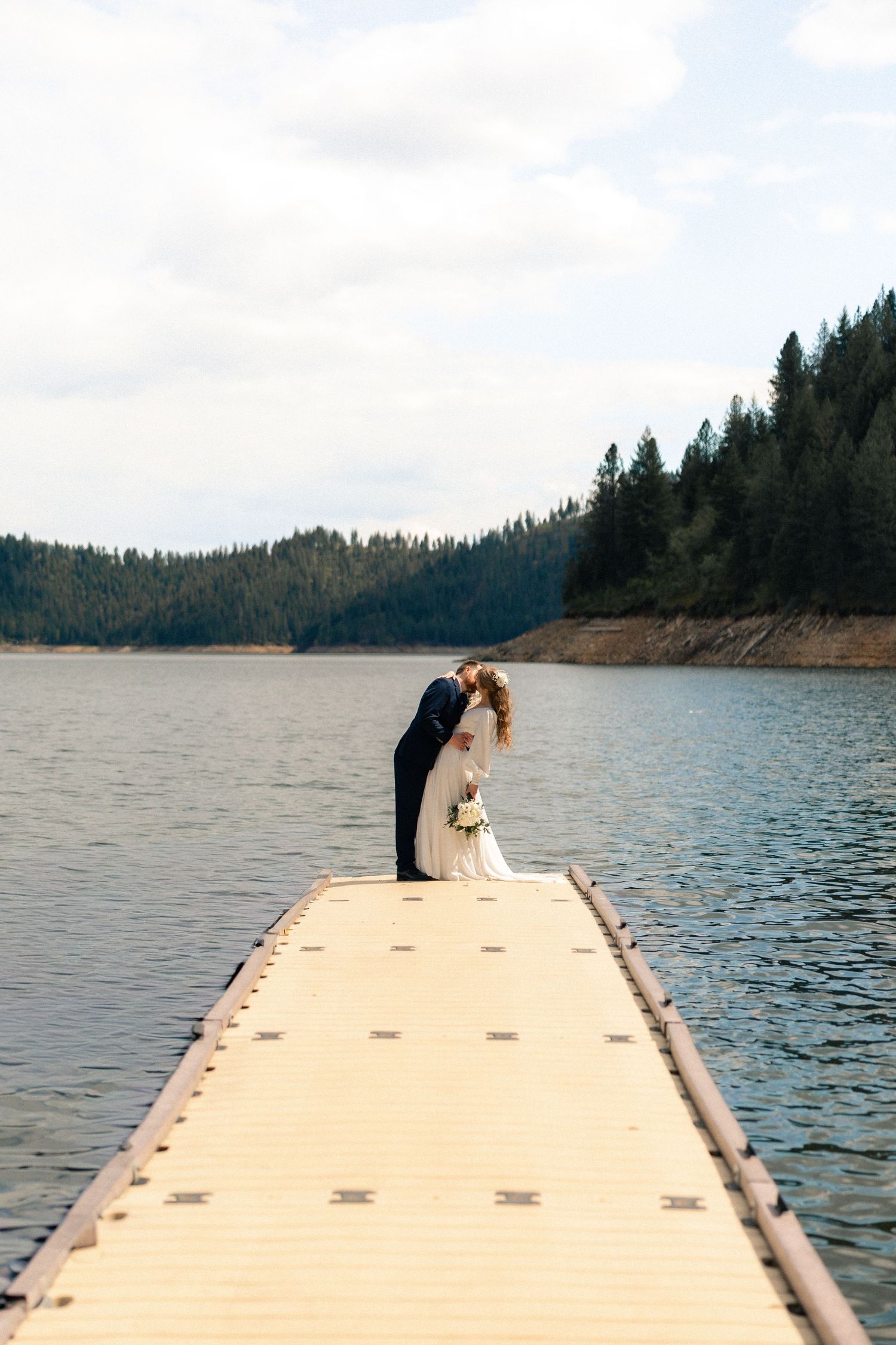 Bride and groom share a romantic kiss on a wooden dock over a calm lake surrounded by evergreen forest.