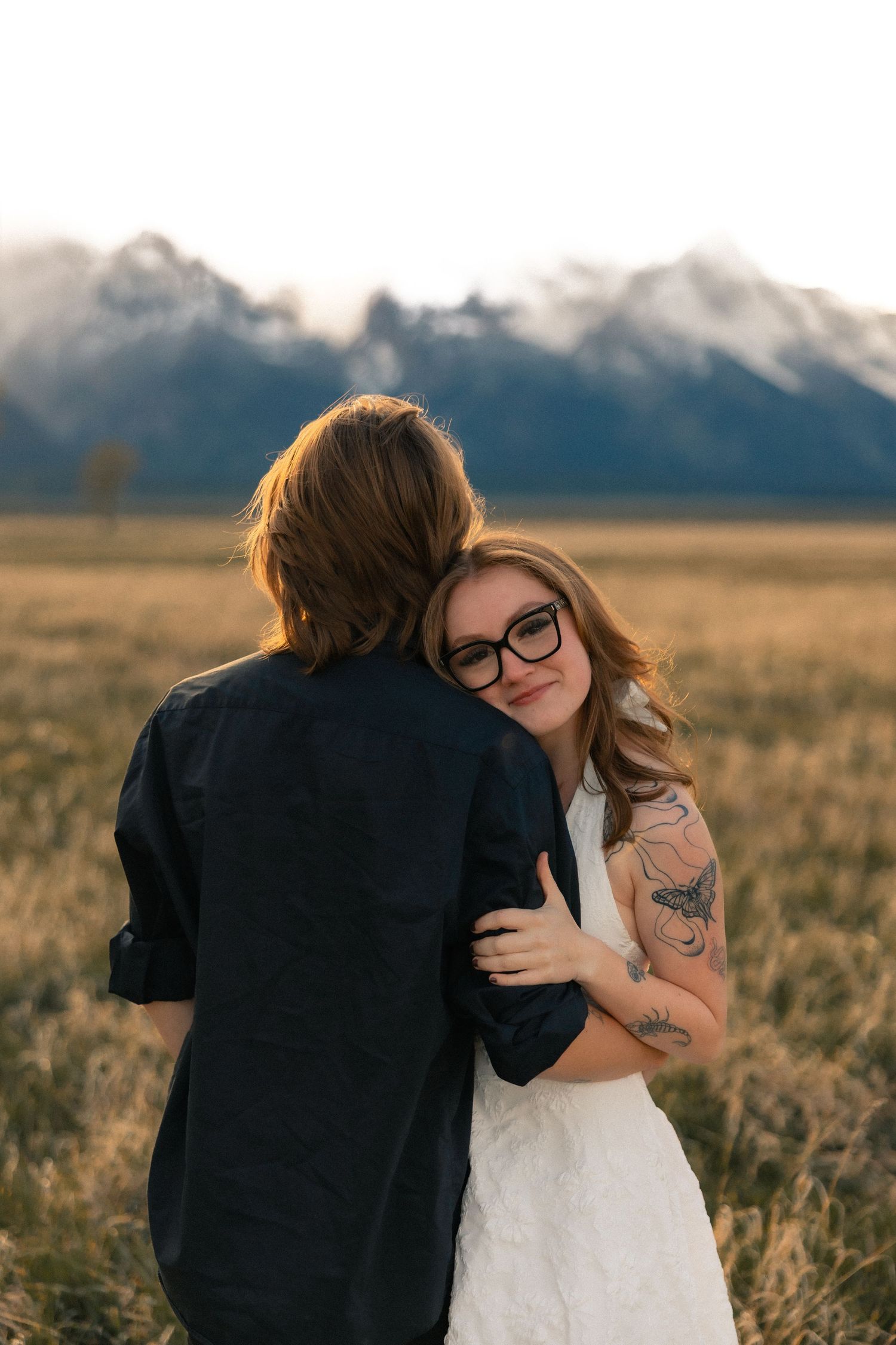 Couple embracing in golden field with snow-capped Grand Teton mountains, bride in white dress with tattoos and glasses.