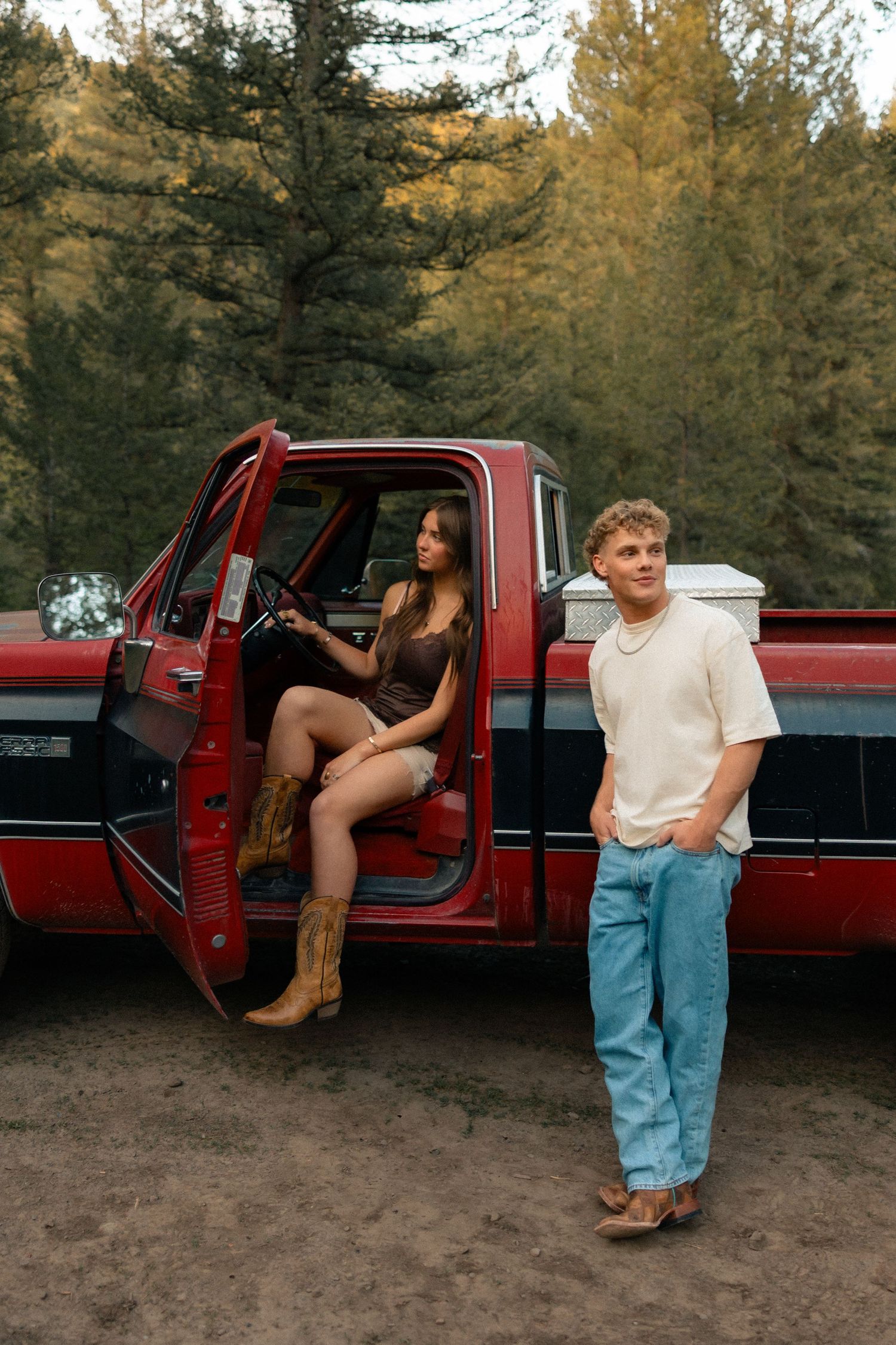 A young couple poses with a vintage red pickup truck surrounded by pine trees, woman in cowboy boots sits in cab.