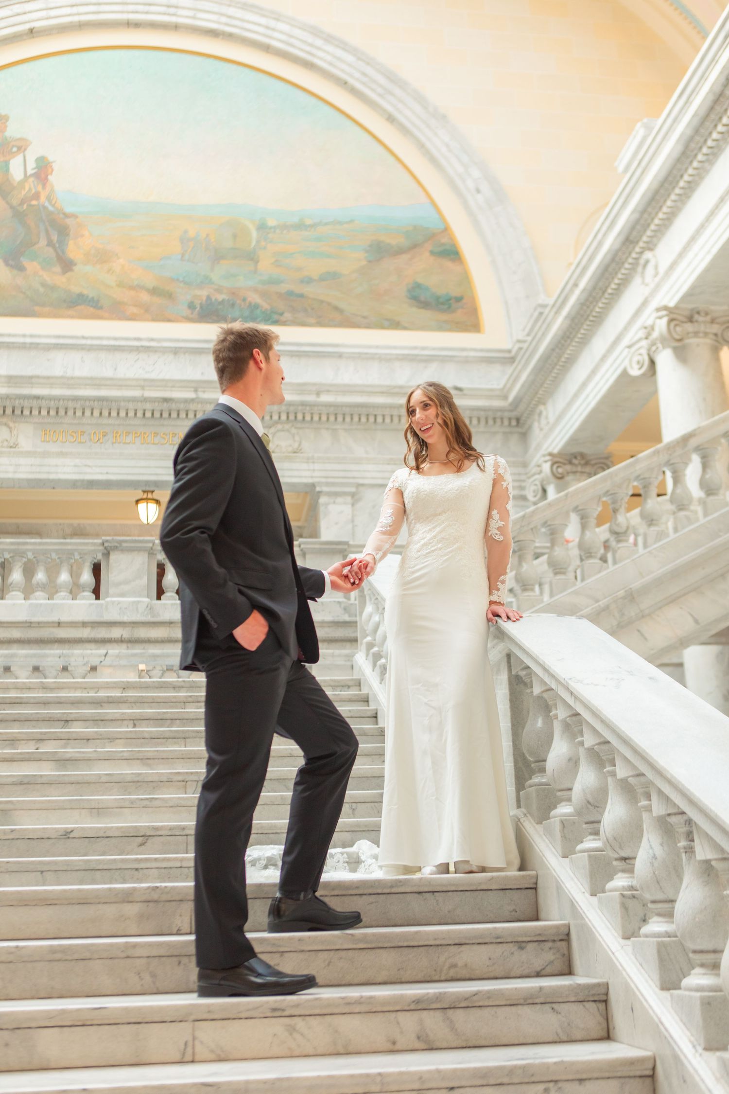 Bride and groom holding hands on marble staircase beneath ornate mural in classic wedding venue.